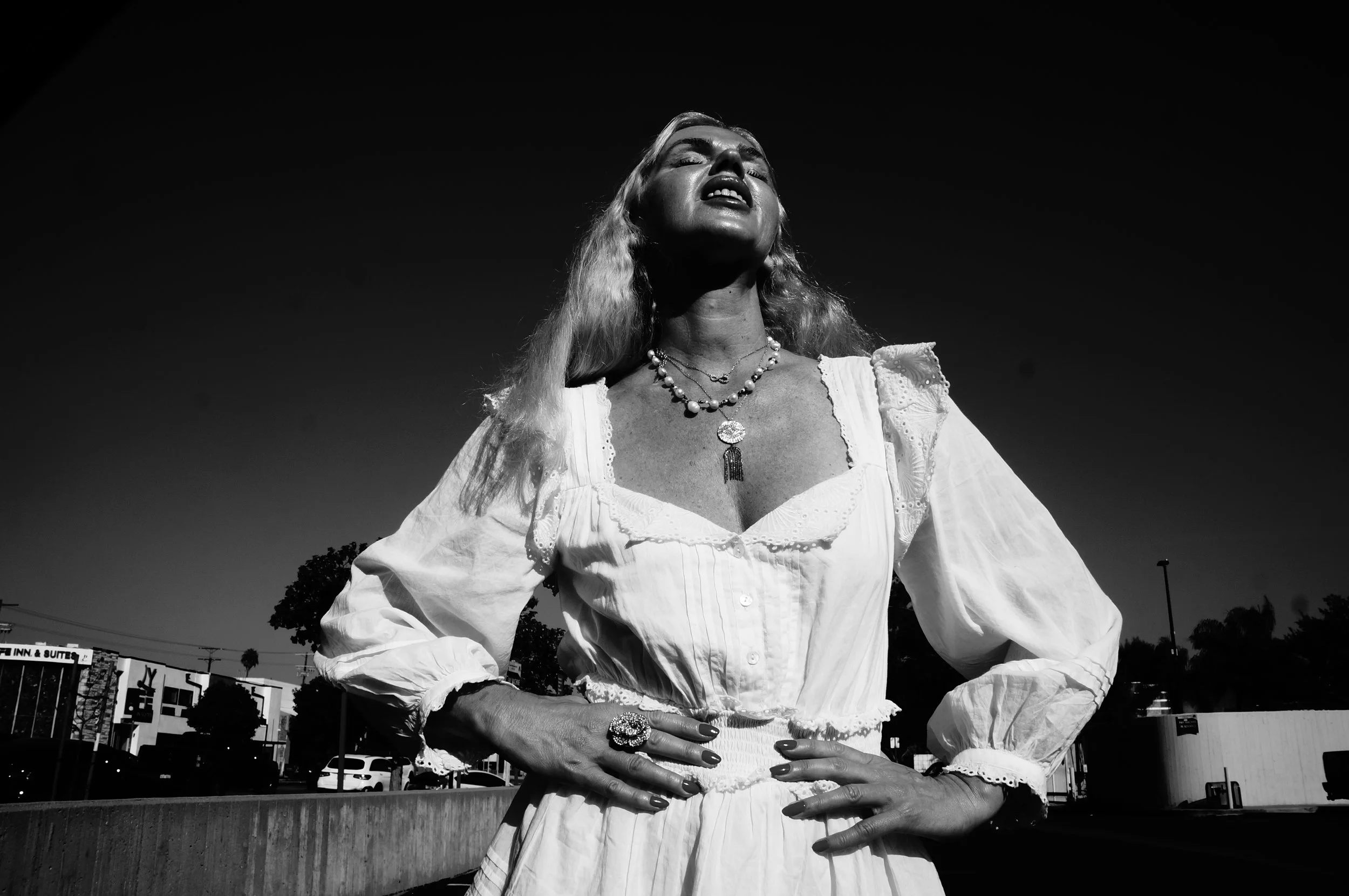 A woman with long, wavy blonde hair, wearing a vintage white dress with puffed sleeves, stands outdoors with her hands on her hips and her head tilted back, sunlit, with a dark sky in the background.
