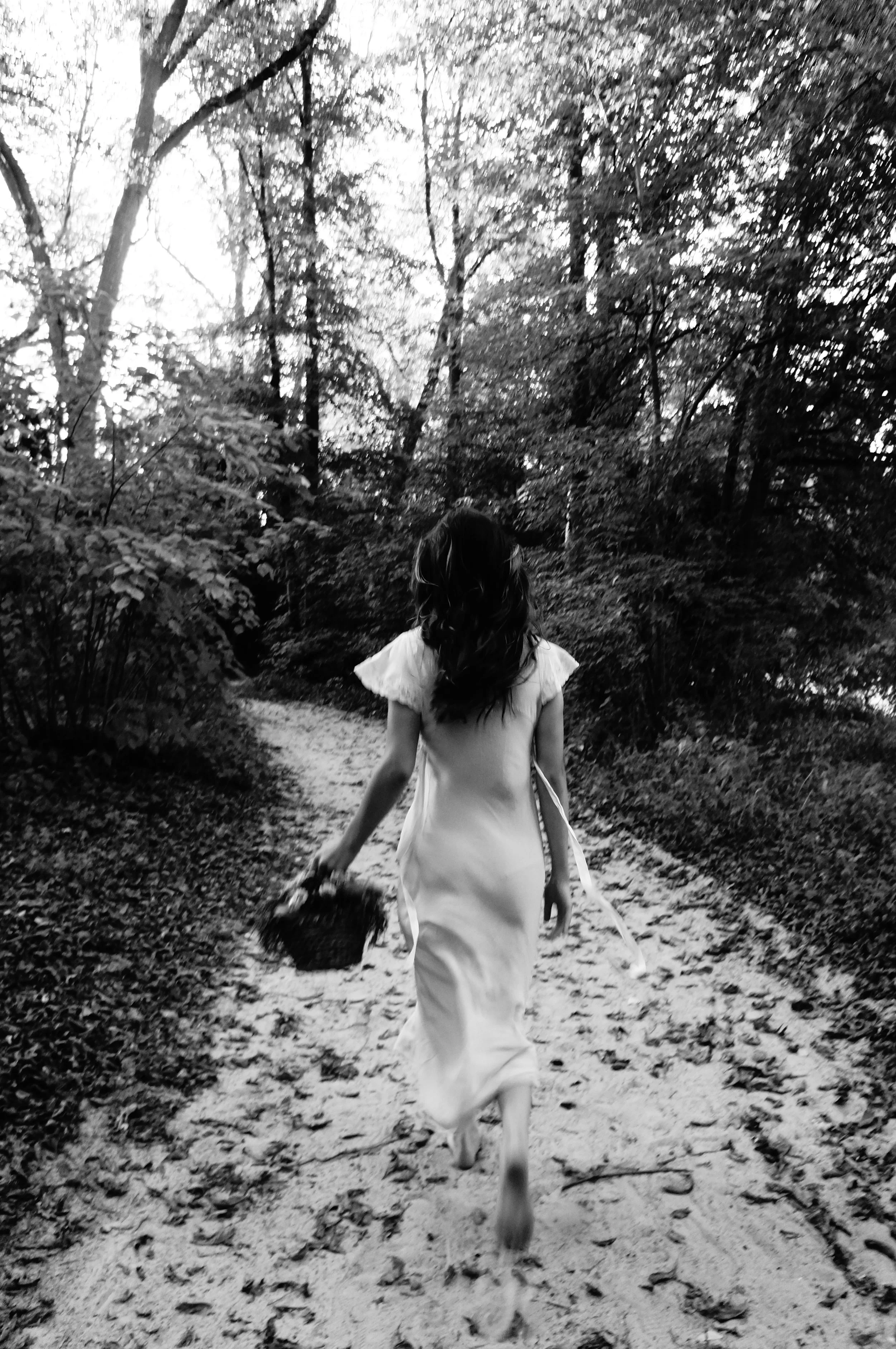 A girl in a light-colored dress walking away on a forest trail, holding a basket, surrounded by trees and foliage.