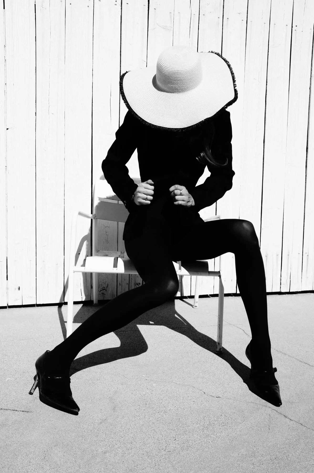 A person dressed in black, sitting on a white bench against a wooden wall, wearing a wide-brimmed hat, black tights, and high heels, with their head bowed and hands clenched.