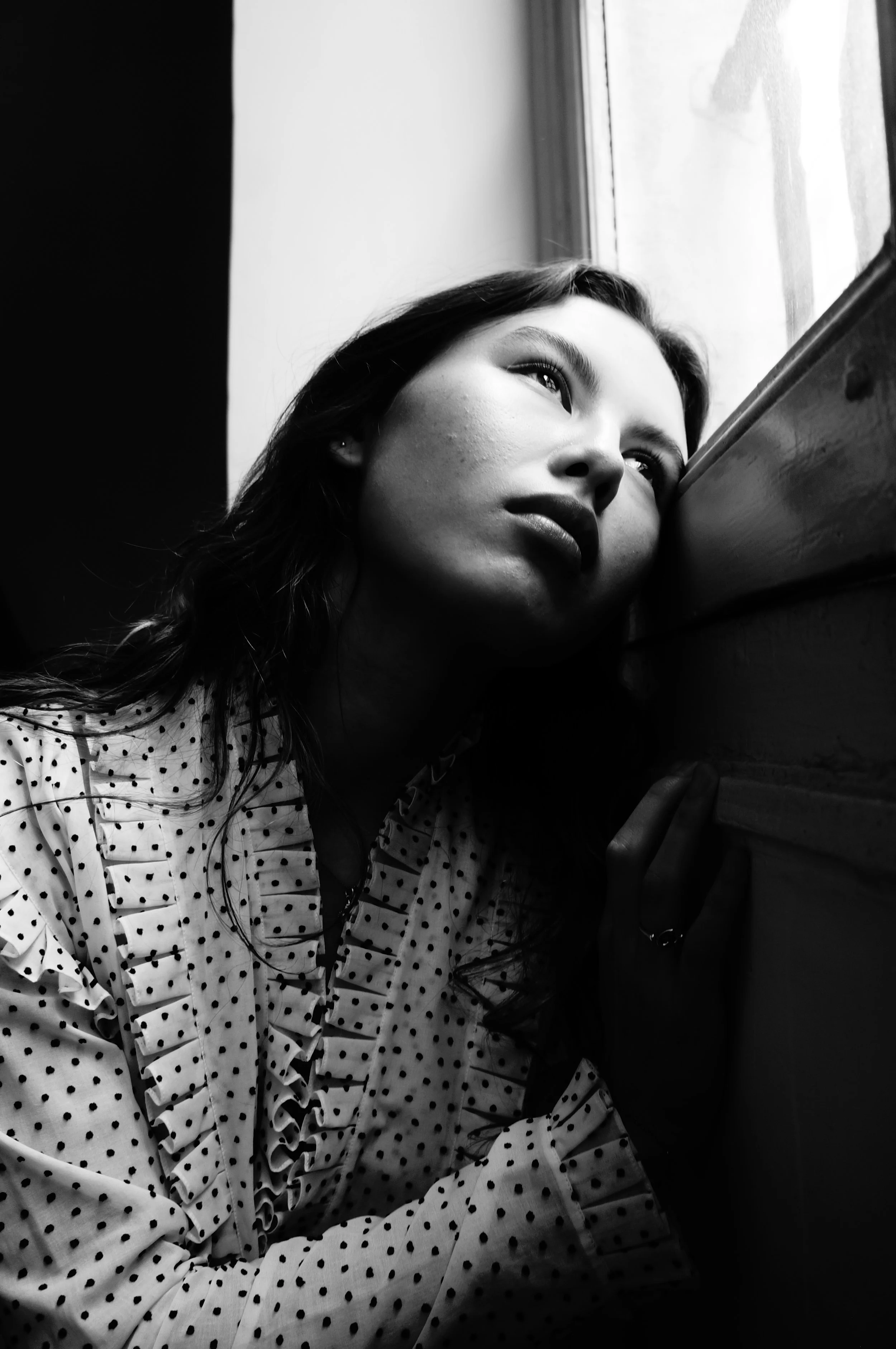 A young woman with wavy hair gazes out the window in black and white, resting her head on her hand.