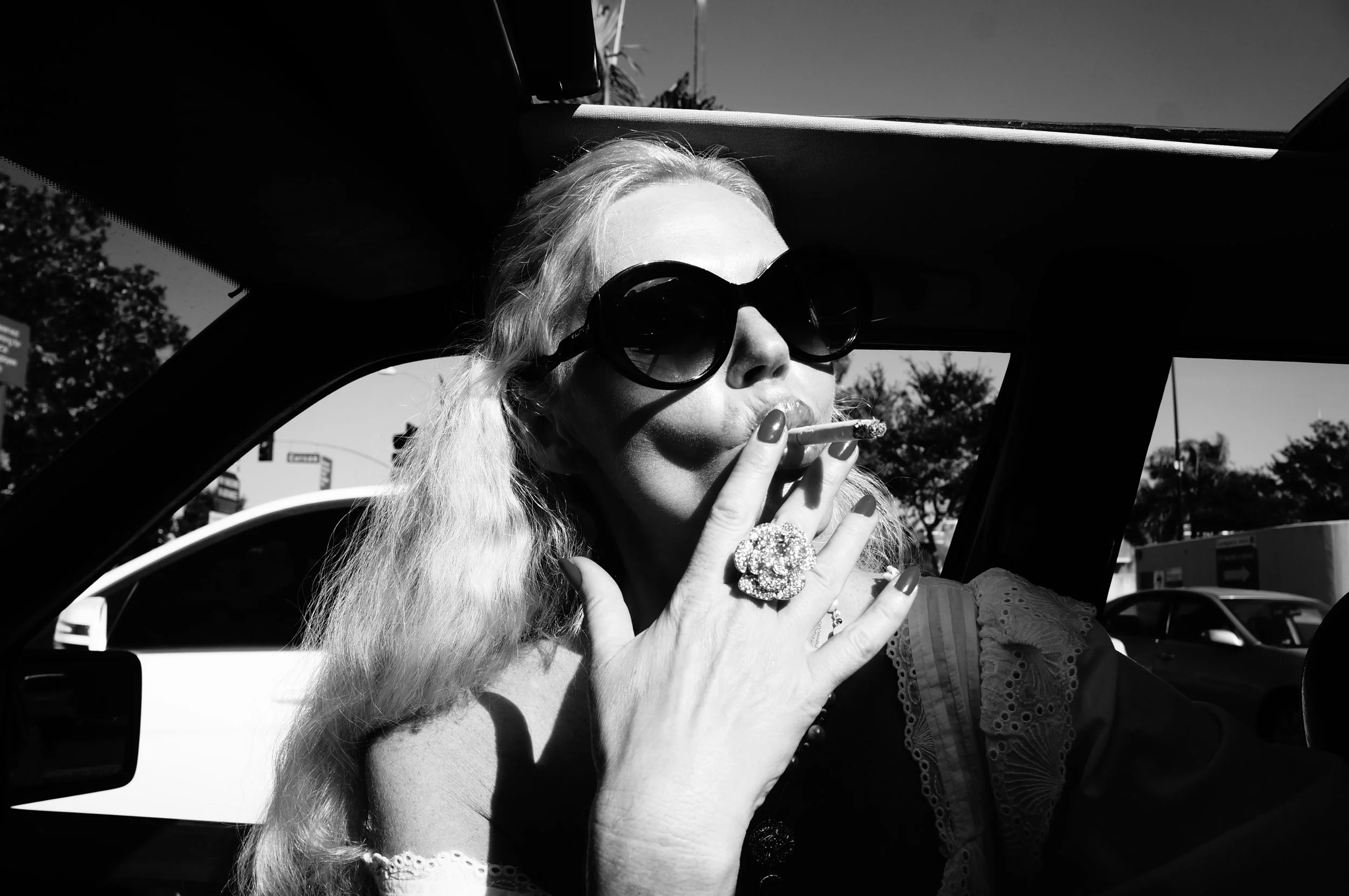 Black and white photo of a woman with long, wavy hair, wearing large sunglasses, smoking a cigarette, and showing a large ring on her finger. She is sitting in a car with the sun shining outside.