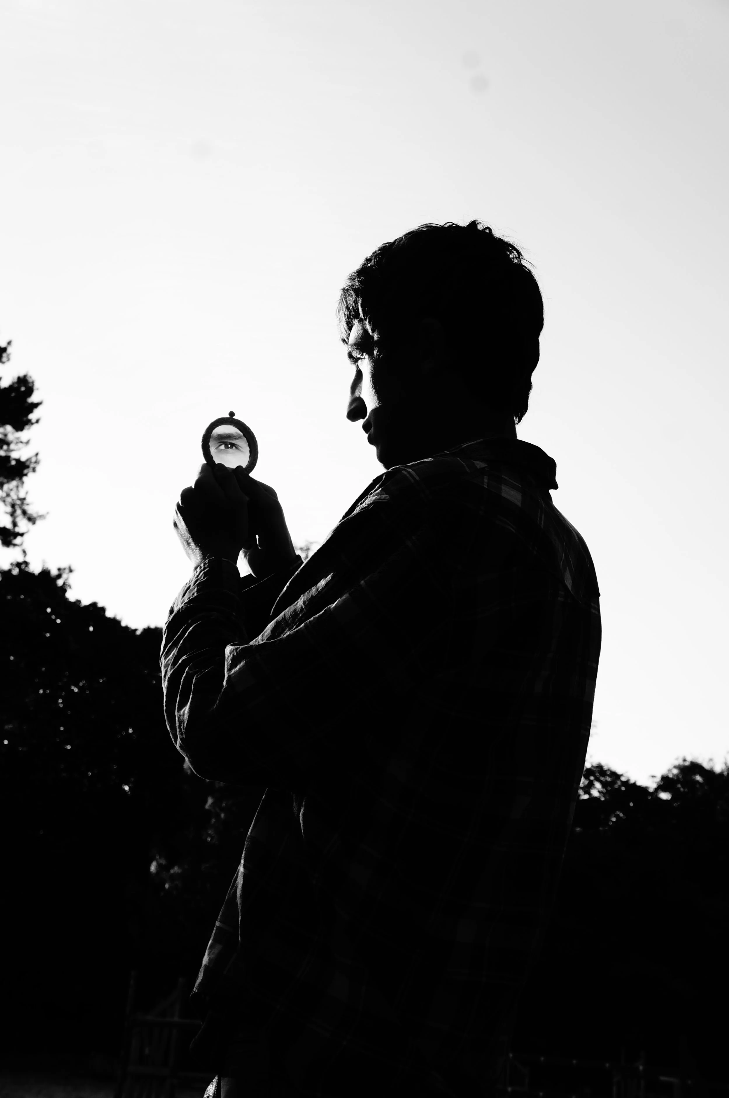 Silhouette of a man holding a small mirror with an eye reflection, outdoors during sunset or sunrise.