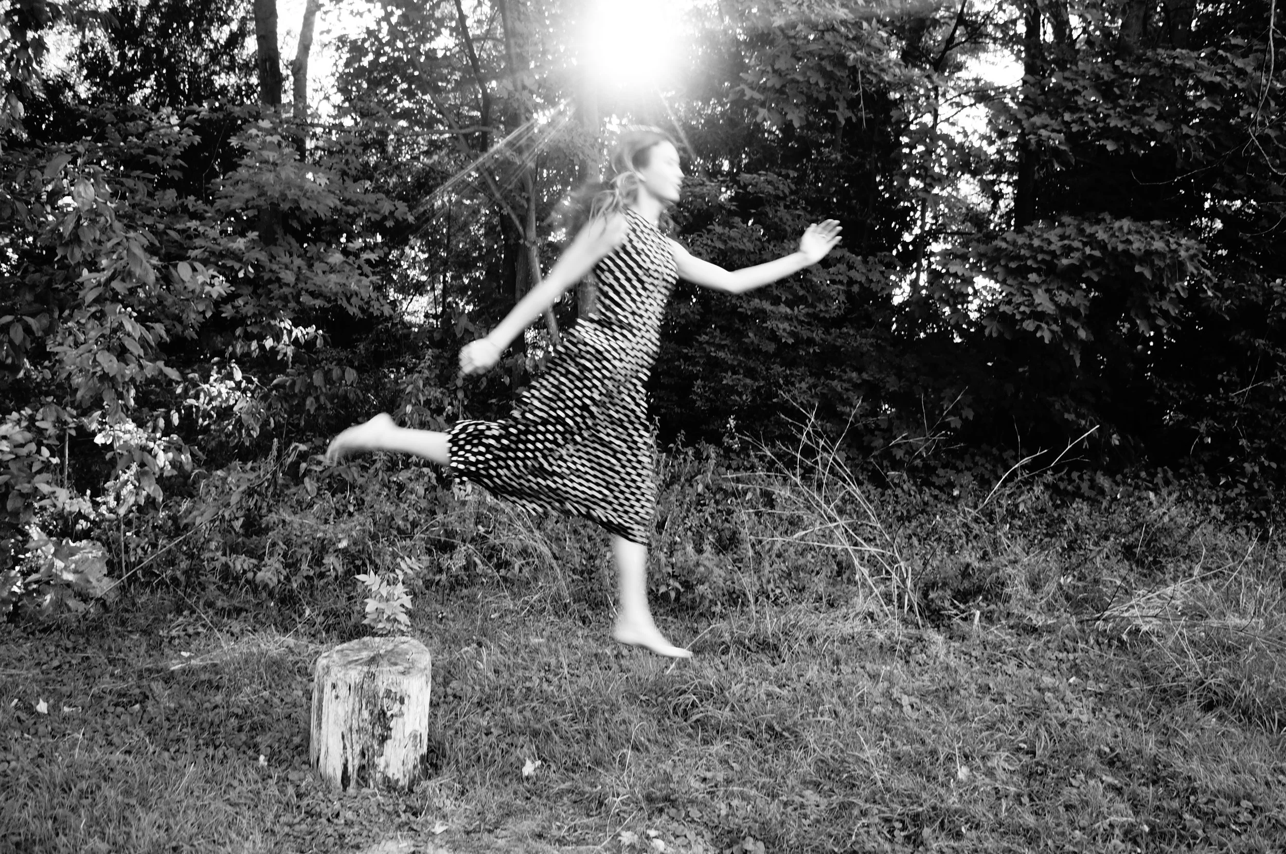 A young girl in a polka dot dress jumping in the air outdoors with trees and bushes in the background, sunlight shining through the trees.