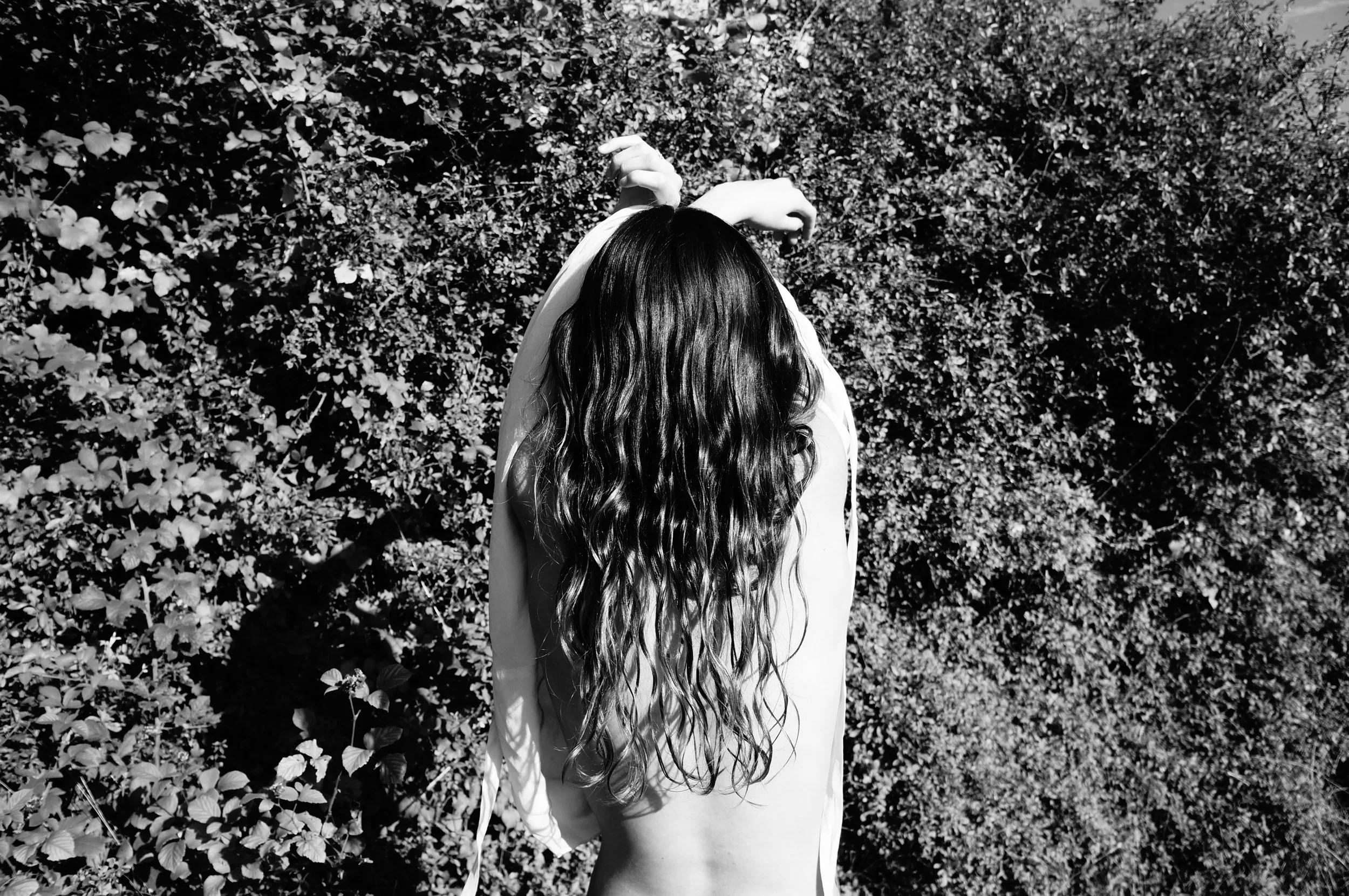 Back view of a woman with long, wavy hair standing outdoors next to a bush.