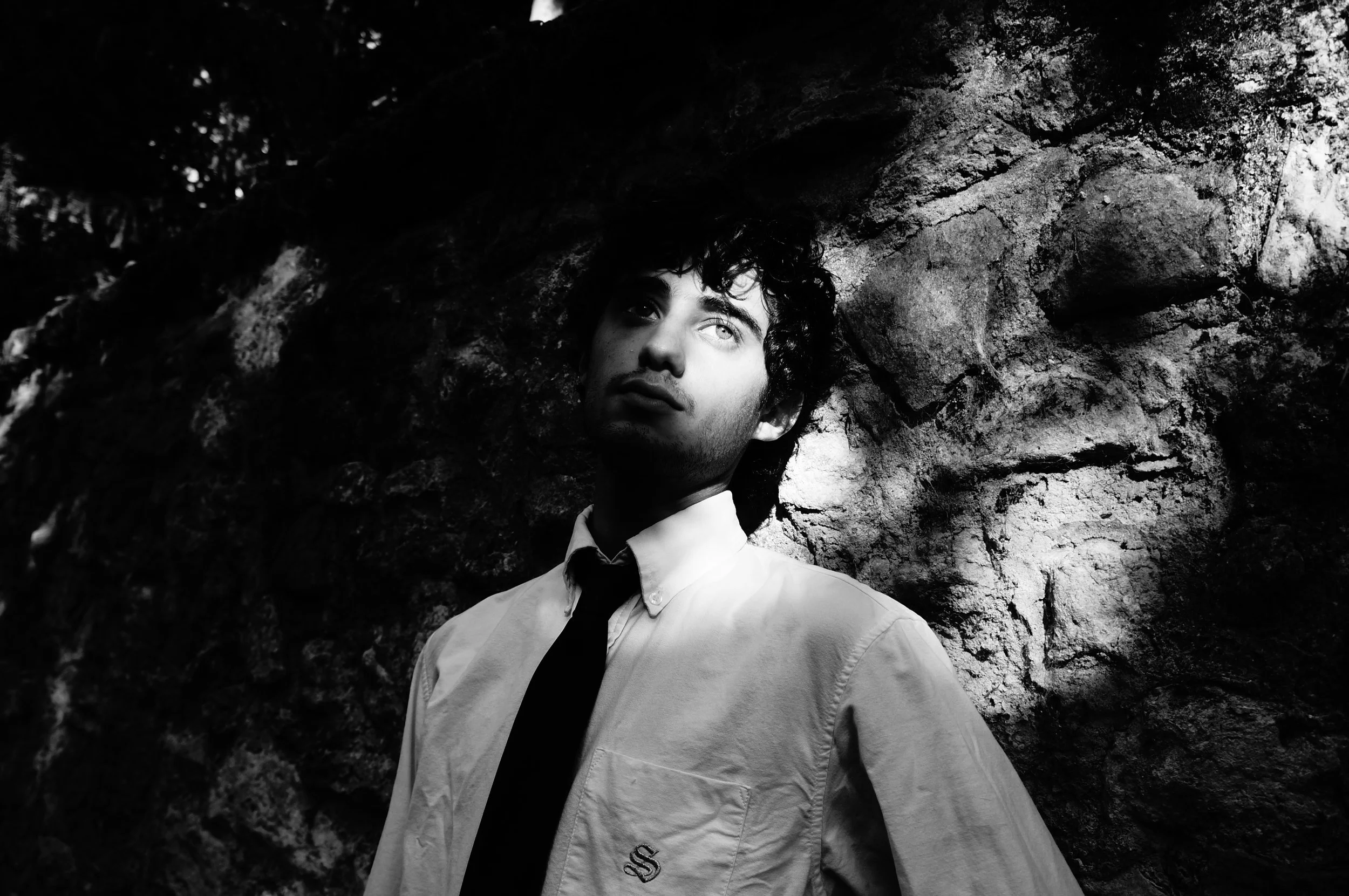 A young man with dark curly hair and light eyes stands outdoors against a rocky background, looking slightly upward, with shadows playing across his face in black and white photography.