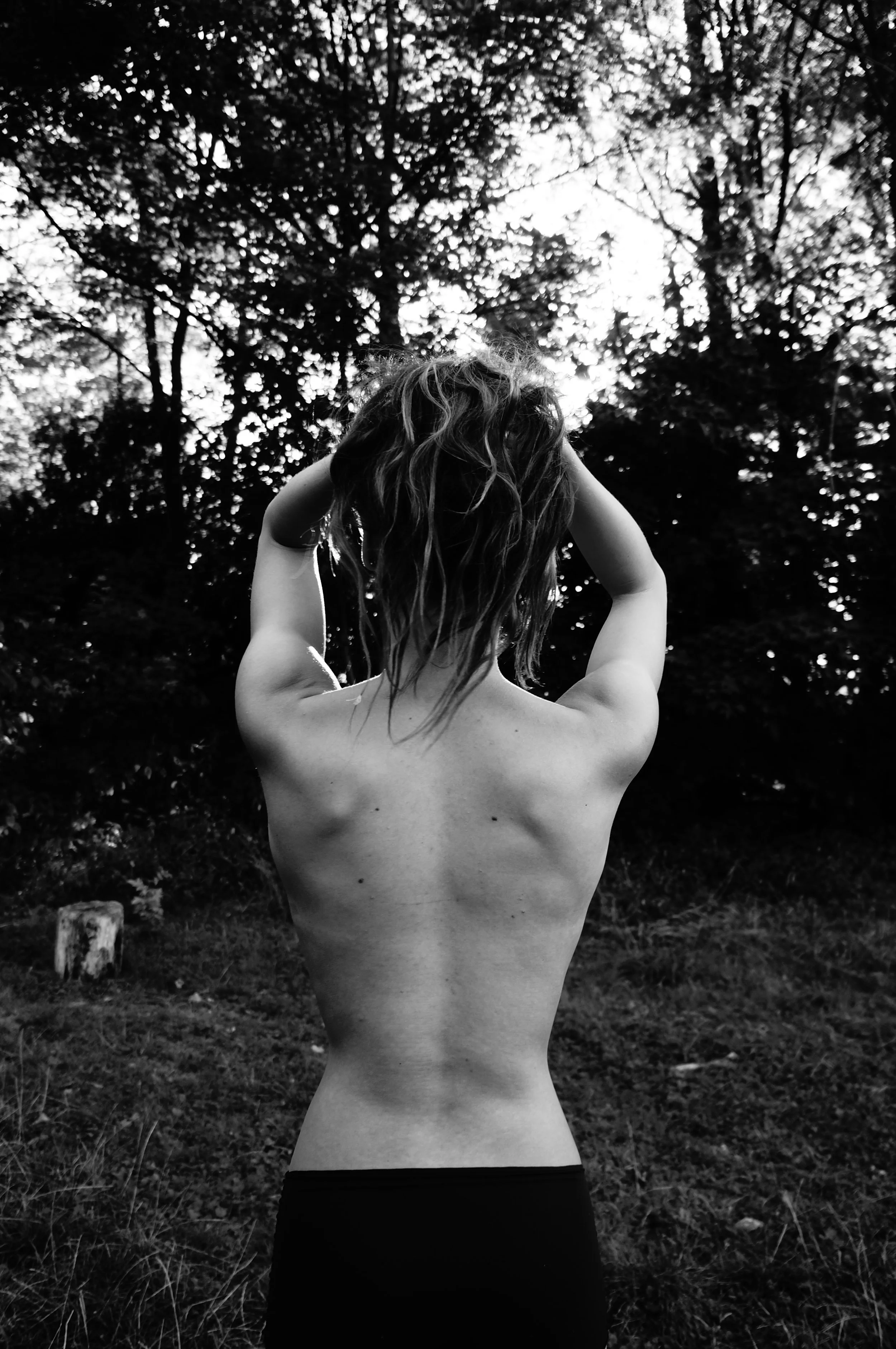 A person with long wavy hair, seen from behind, standing outdoors with arms raised behind their head, surrounded by trees, in black and white photograph.