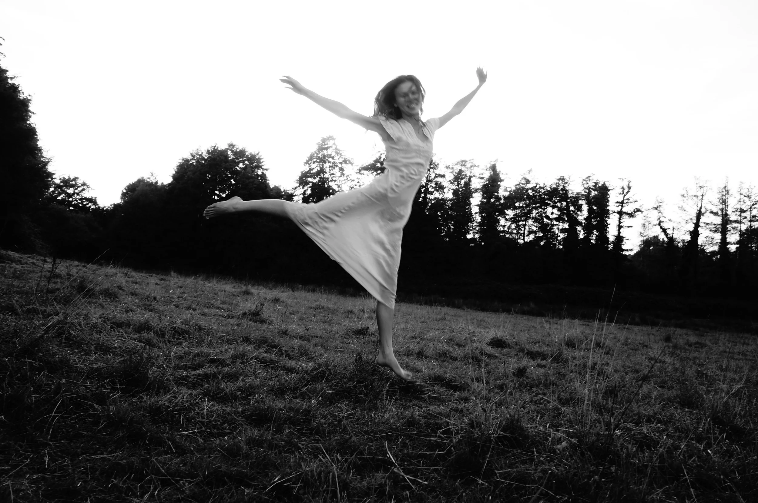 A woman in a dress joyfully jumping in an open field with trees in the background, captured in black and white.