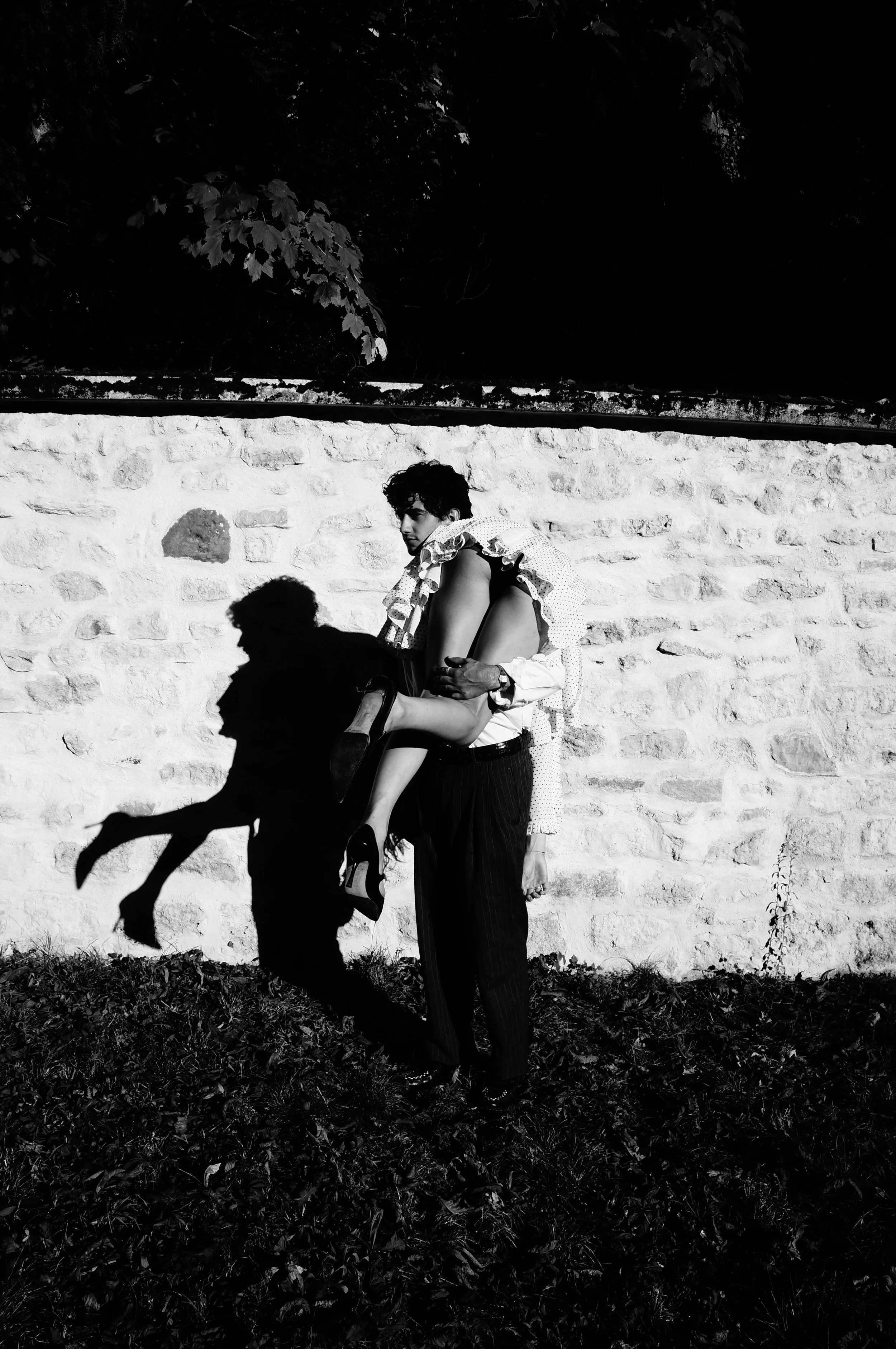 A black and white photo of a man standing against a stone wall, carrying a woman in his arms with her legs hanging down. The woman is dressed in high heels and a patterned outfit, while the man is wearing dark pants and a patterned shirt. Their shado