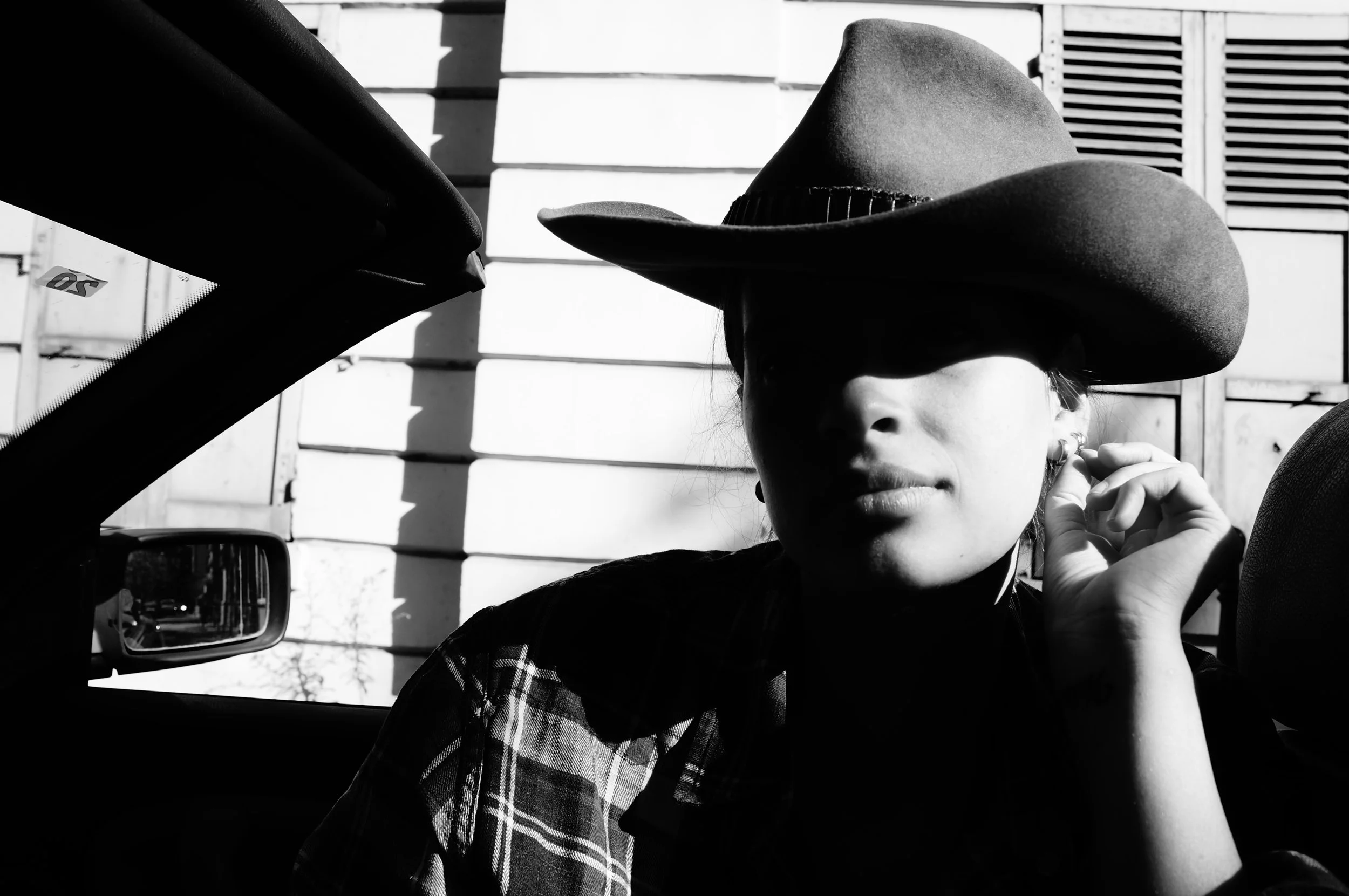 Black and white photo of a woman sitting in a convertible car, wearing a wide brim cowboy hat and a plaid shirt, with her hand near her ear, in front of a wooden wall.