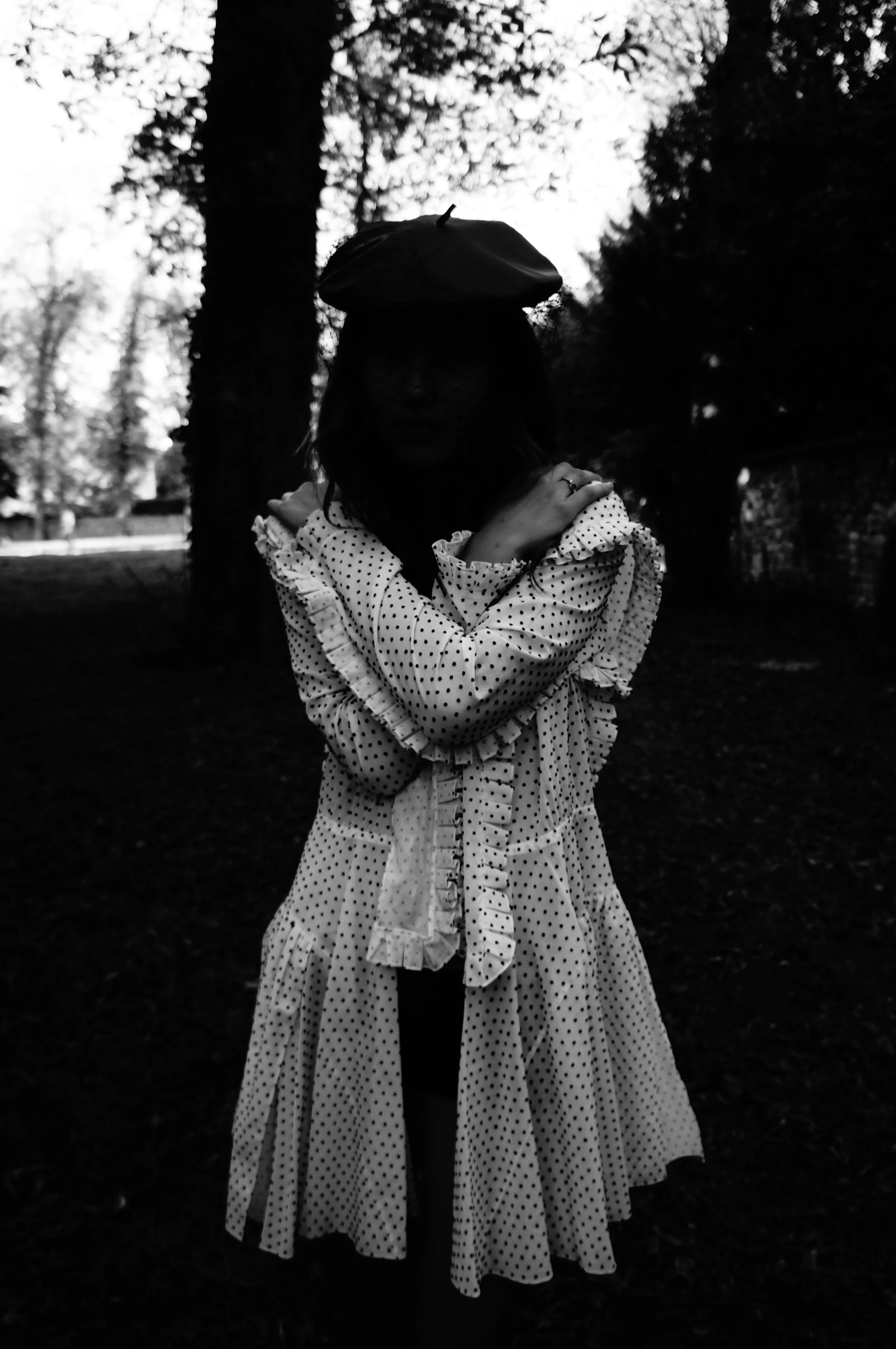 Silhouette of a woman wearing a polka dot dress and a hat, standing outdoors among trees at dusk or dawn.
