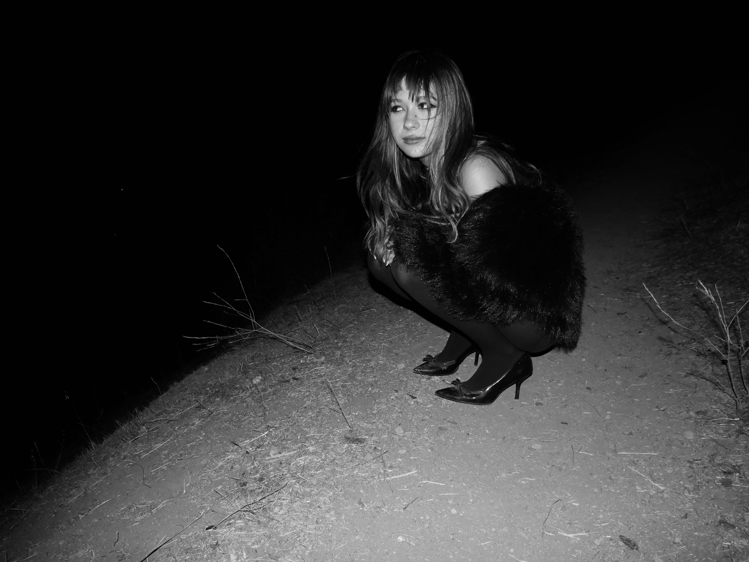 A woman crouching outdoors at night, dressed in high heels, a fur coat, and dark clothing, with a dark background.