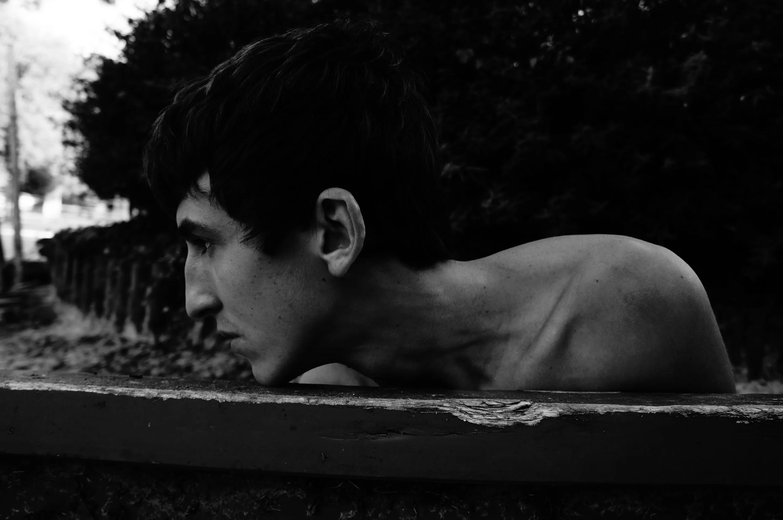 Black and white photo of a young man with dark hair leaning forward on a wooden surface outdoors, with trees in the background.