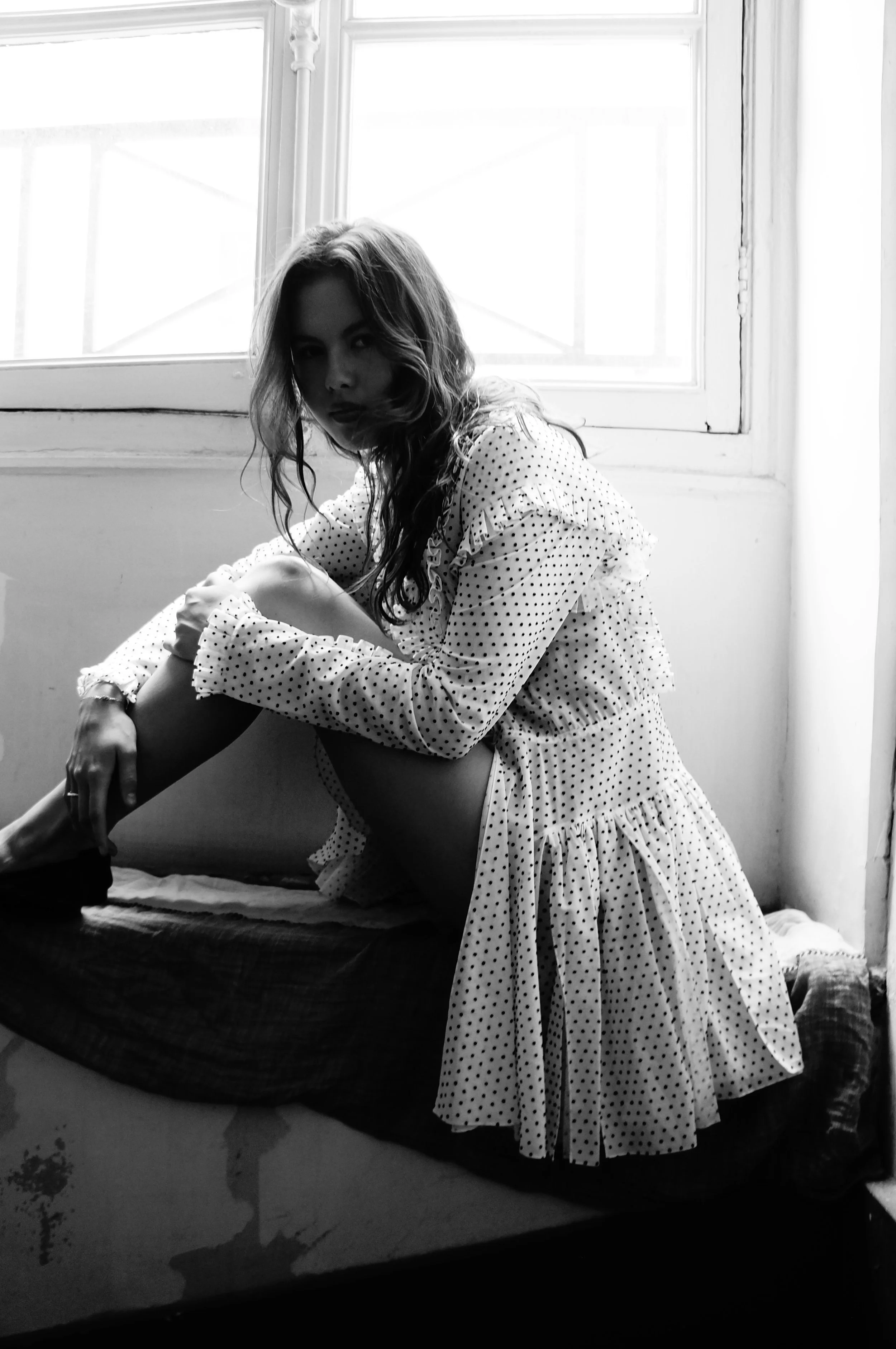 A black-and-white photo of a woman with long hair sitting on a windowsill, wearing a polka dot dress. She is sitting with one leg bent and the other extended, looking directly at the camera.