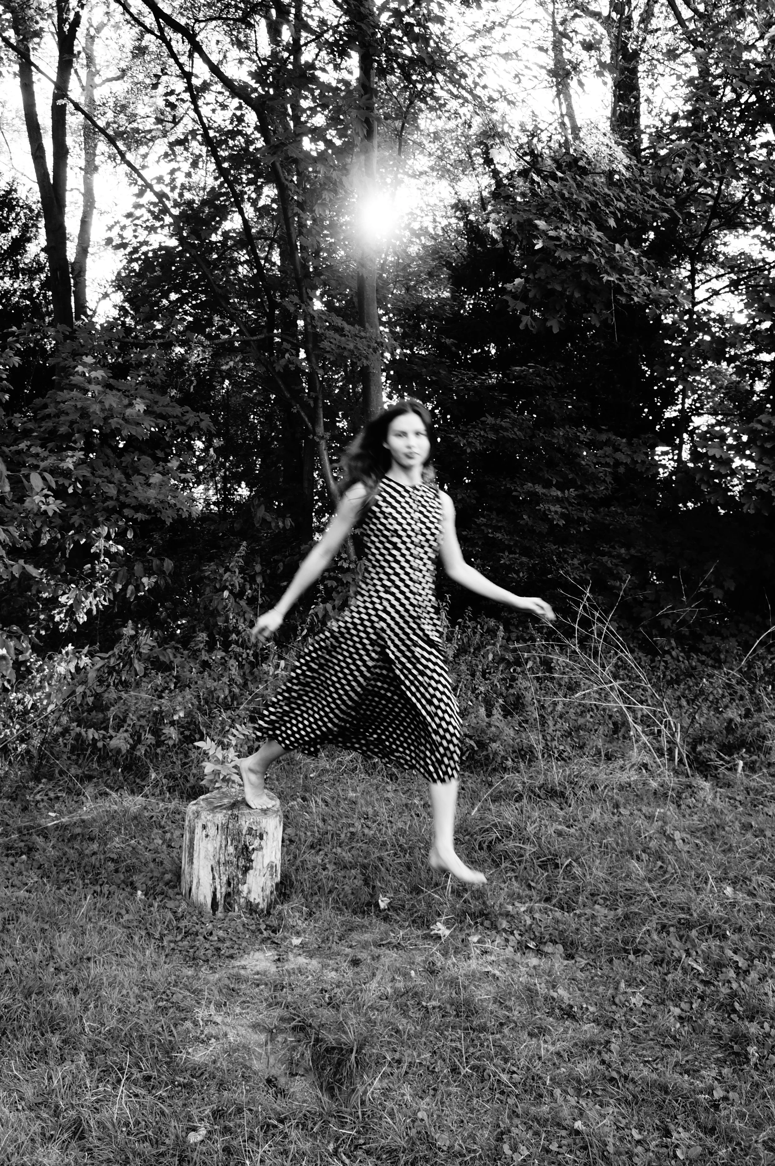 A young woman with long dark hair in a patterned dress balancing on a tree stump in a wooded area with sunlight filtering through the trees in black and white.