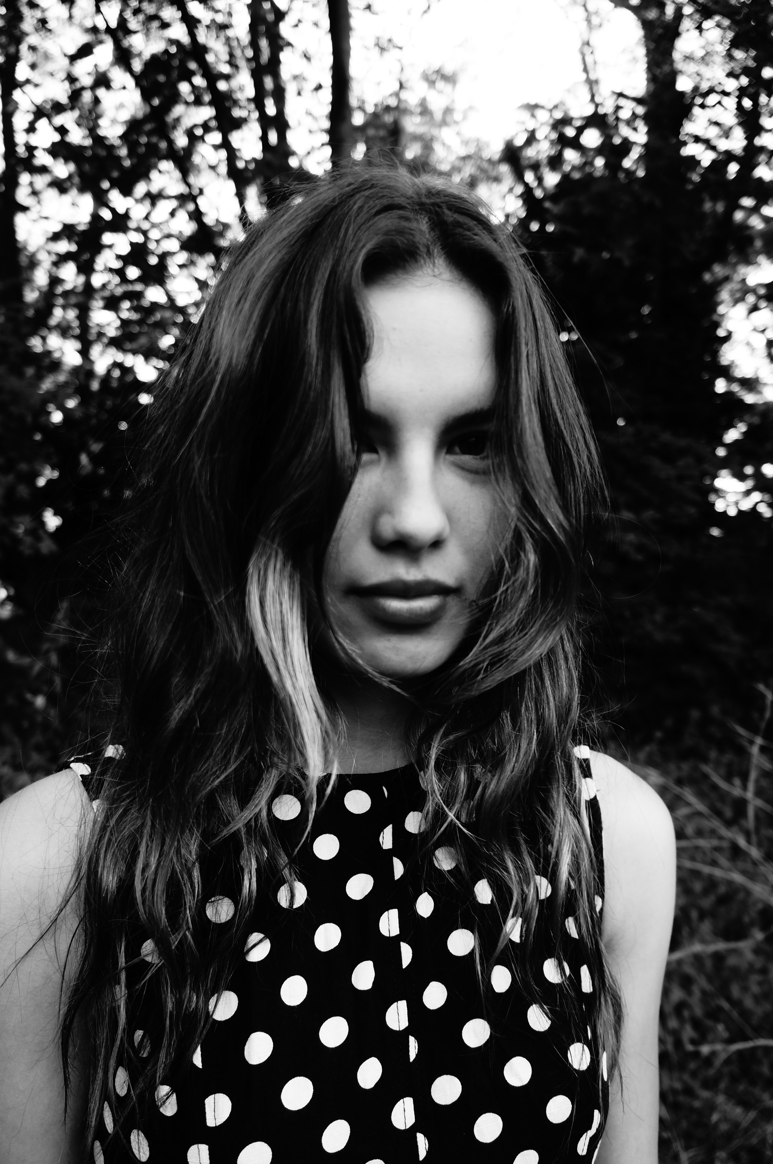 A black and white photo of a young woman with wavy hair, wearing a sleeveless polka dot top, standing outdoors with trees in the background.