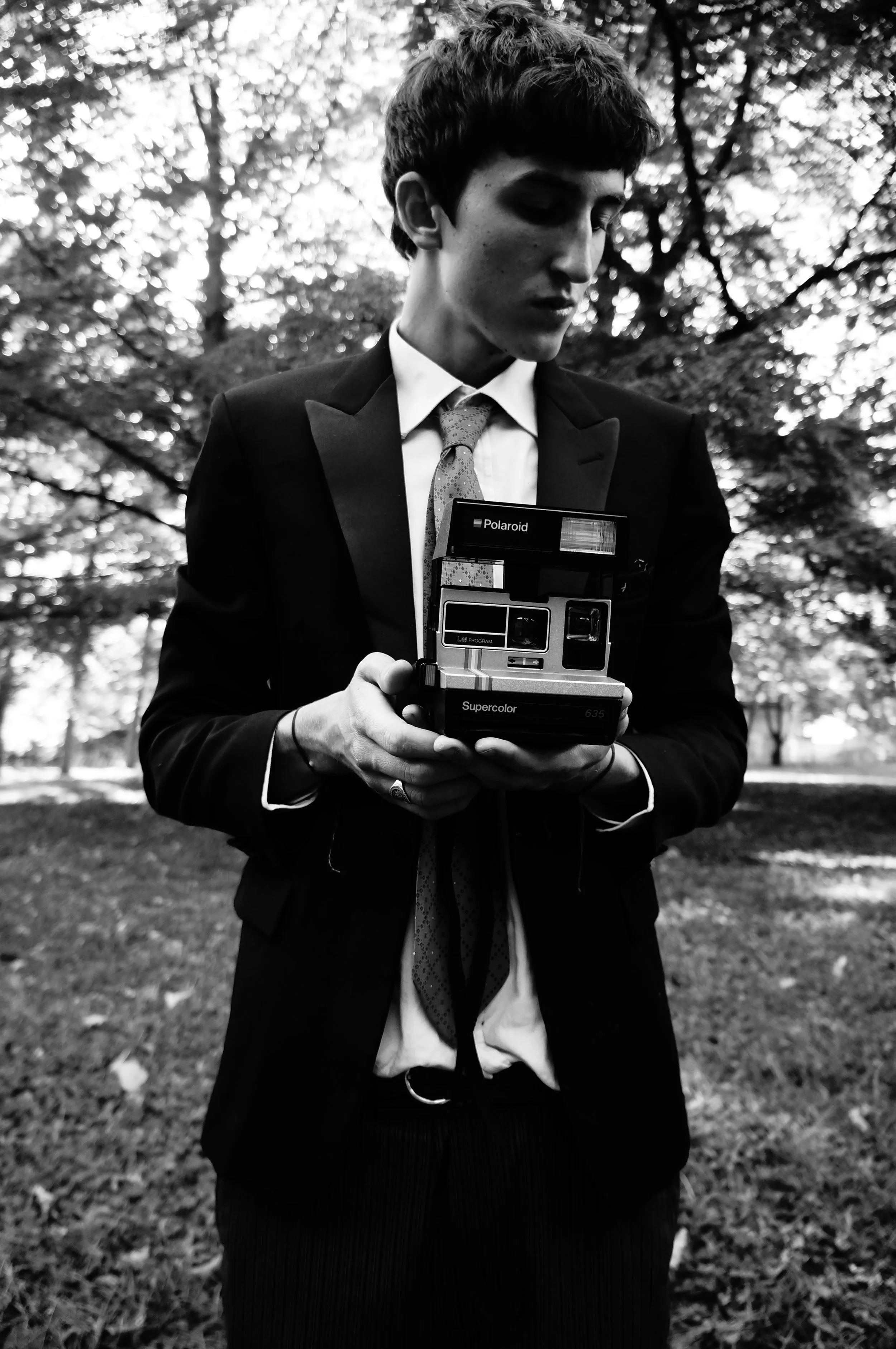 A young man in a suit and tie holding a vintage Polaroid camera outdoors in a park with trees in the background.