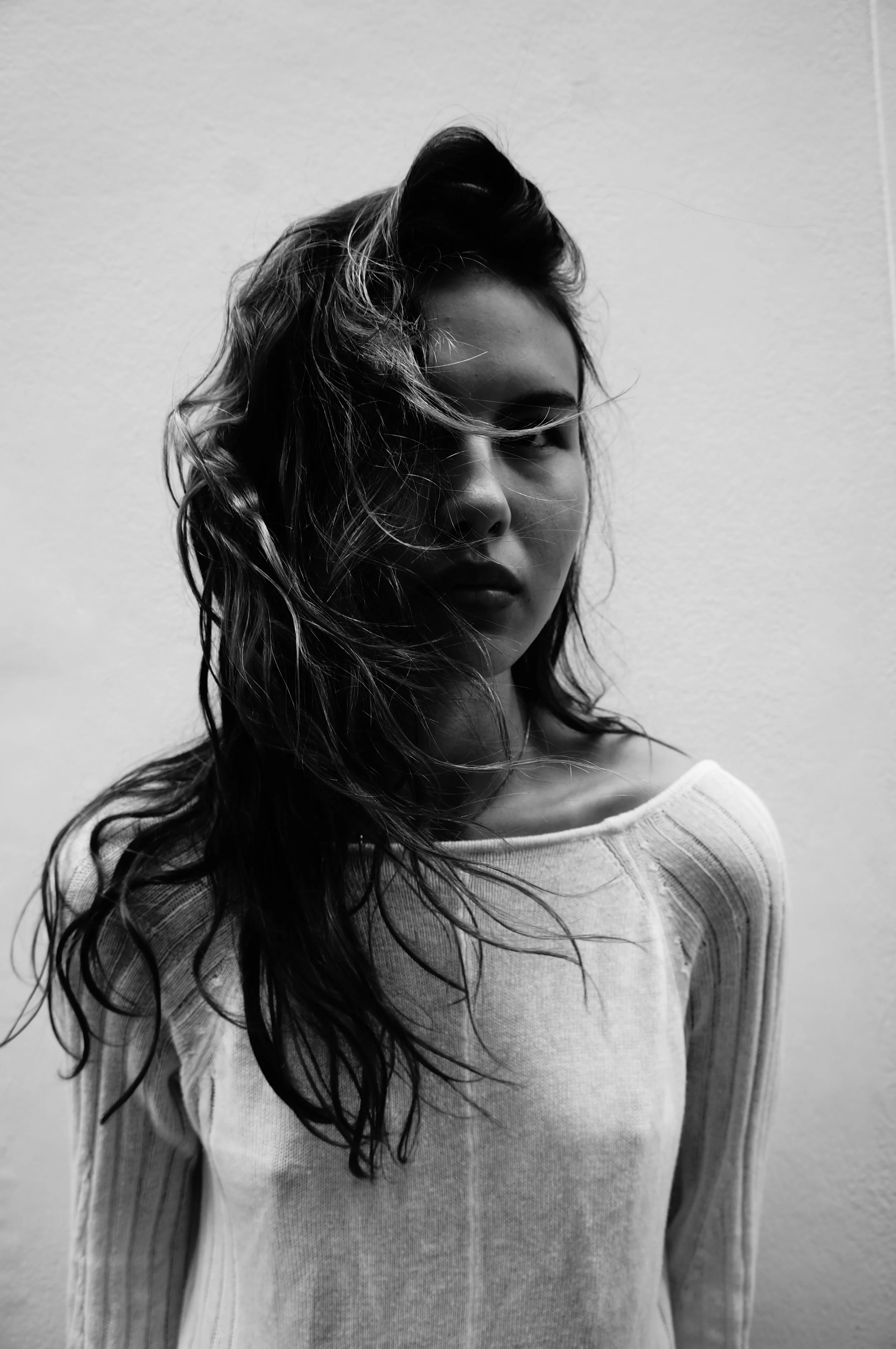 Black and white photo of a woman with long, wavy hair partially covering her face, standing against a plain wall.