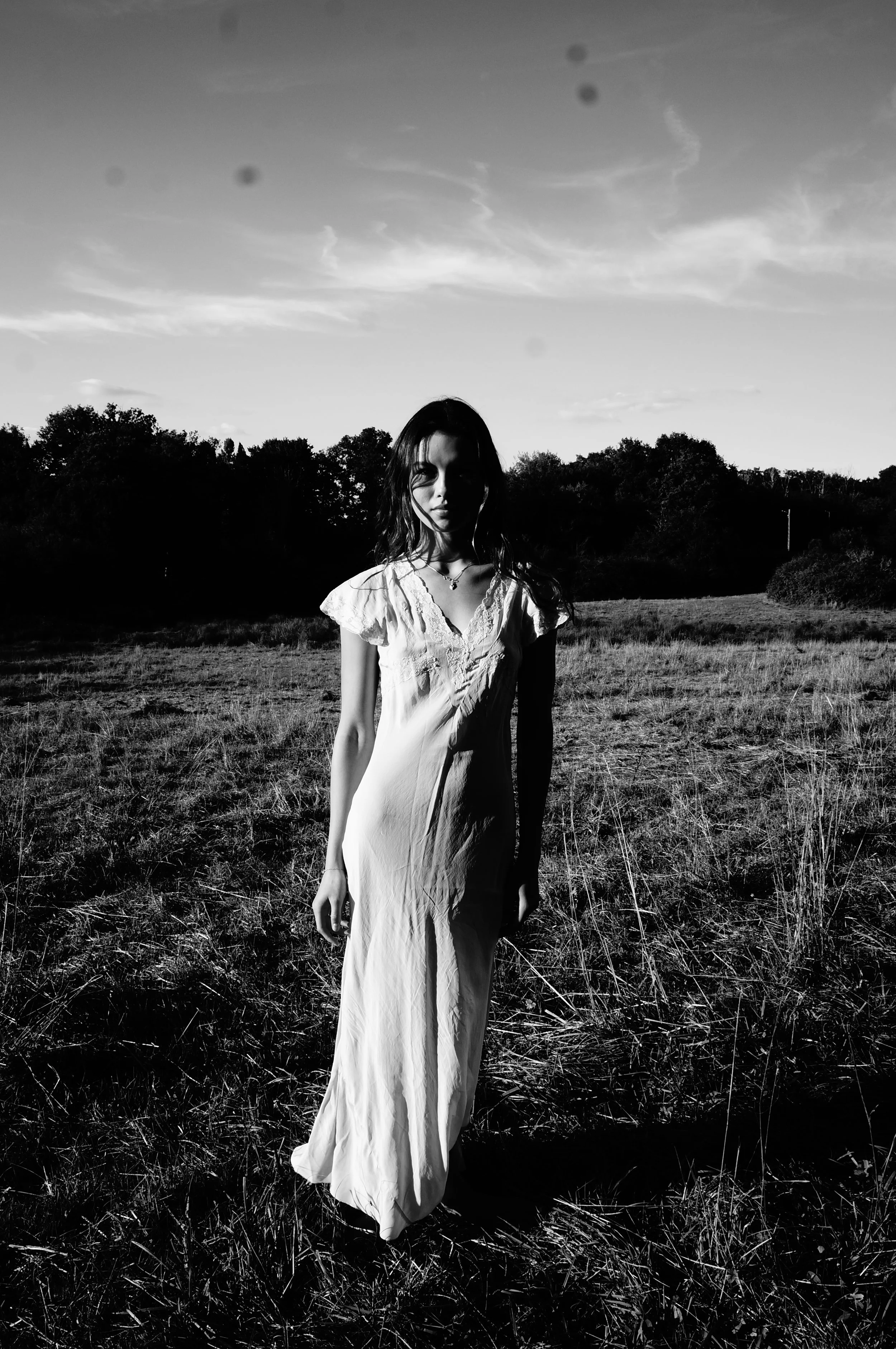 A woman in a long dress standing in a grassy field with trees in the background, captured in black and white.