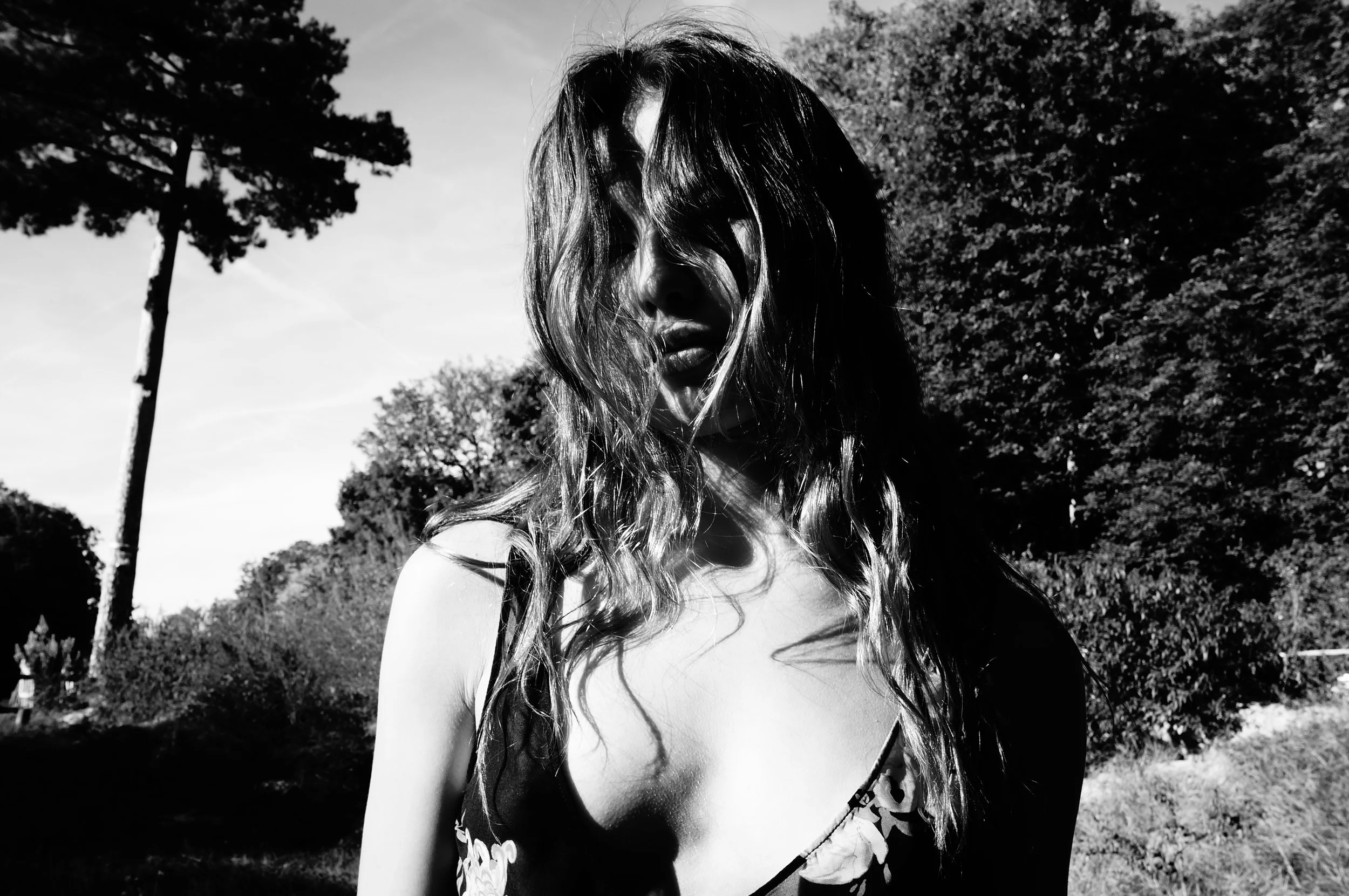 A black and white photo of a woman outdoors, with trees and sky in the background. Her face is partially obscured by her hair, and she is wearing a sleeveless top.