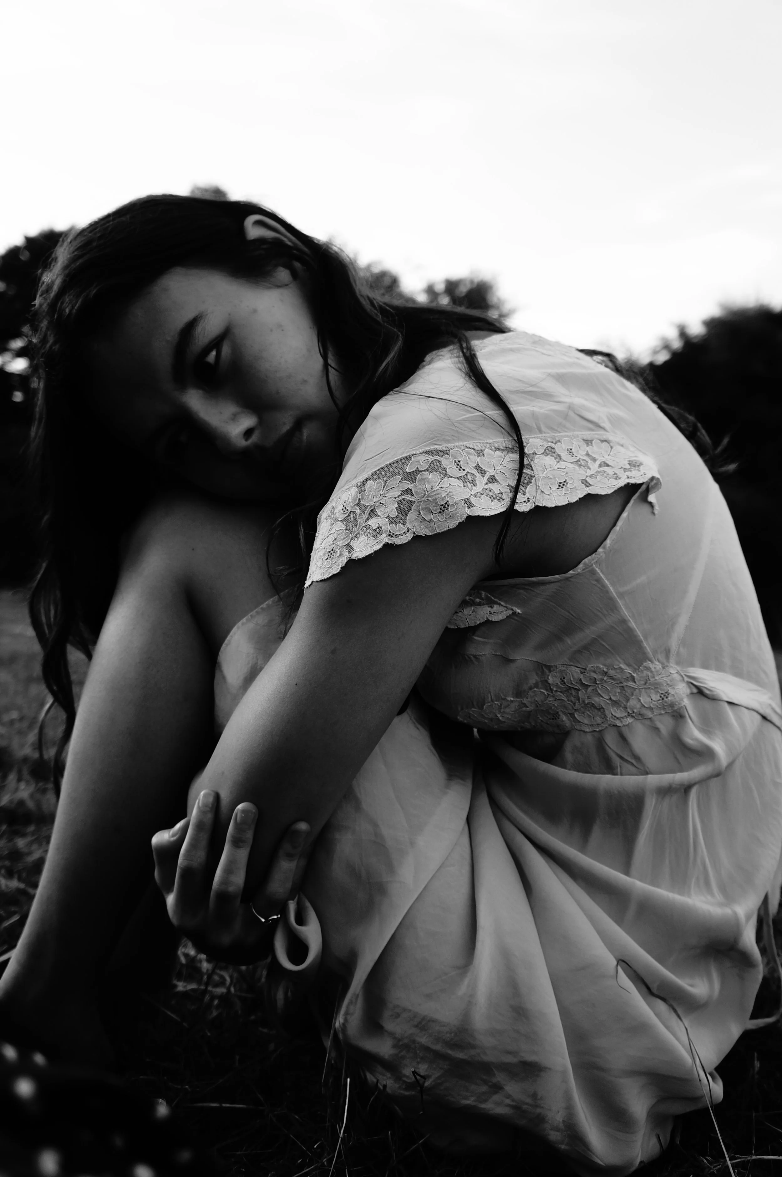 Black-and-white photo of a woman with long dark hair, wearing a dress with lace details, sitting outdoors, looking directly at the camera with a pensive expression, surrounded by trees and sky in the background.