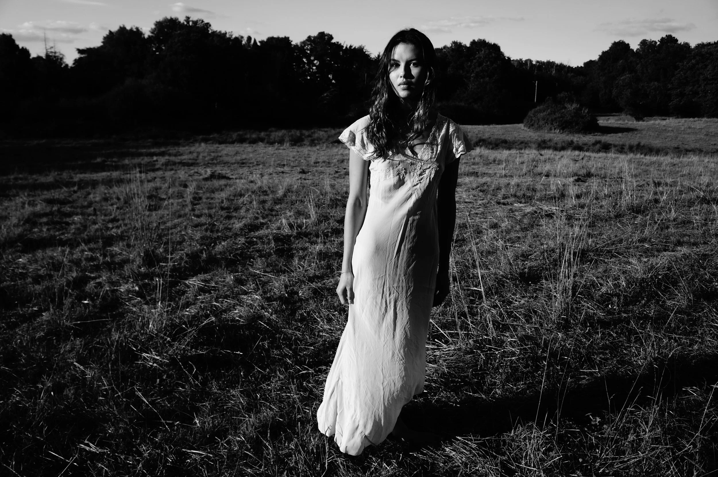A young woman with long hair wearing a light-colored dress standing alone in an outdoor grassy field with trees and sky in the background, captured in black and white.