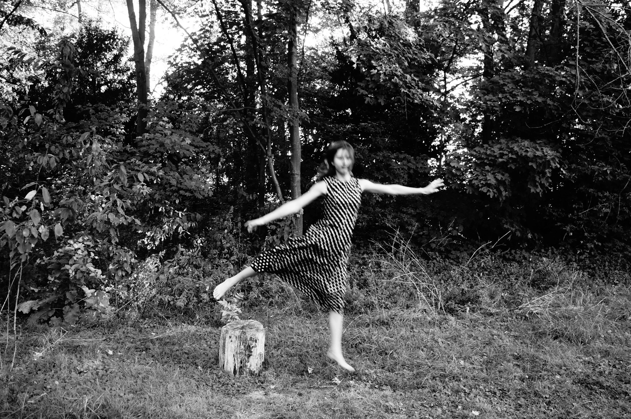 A young girl in a patterned dress balancing on a wooden stump in a wooded outdoor setting, with trees and foliage in the background.