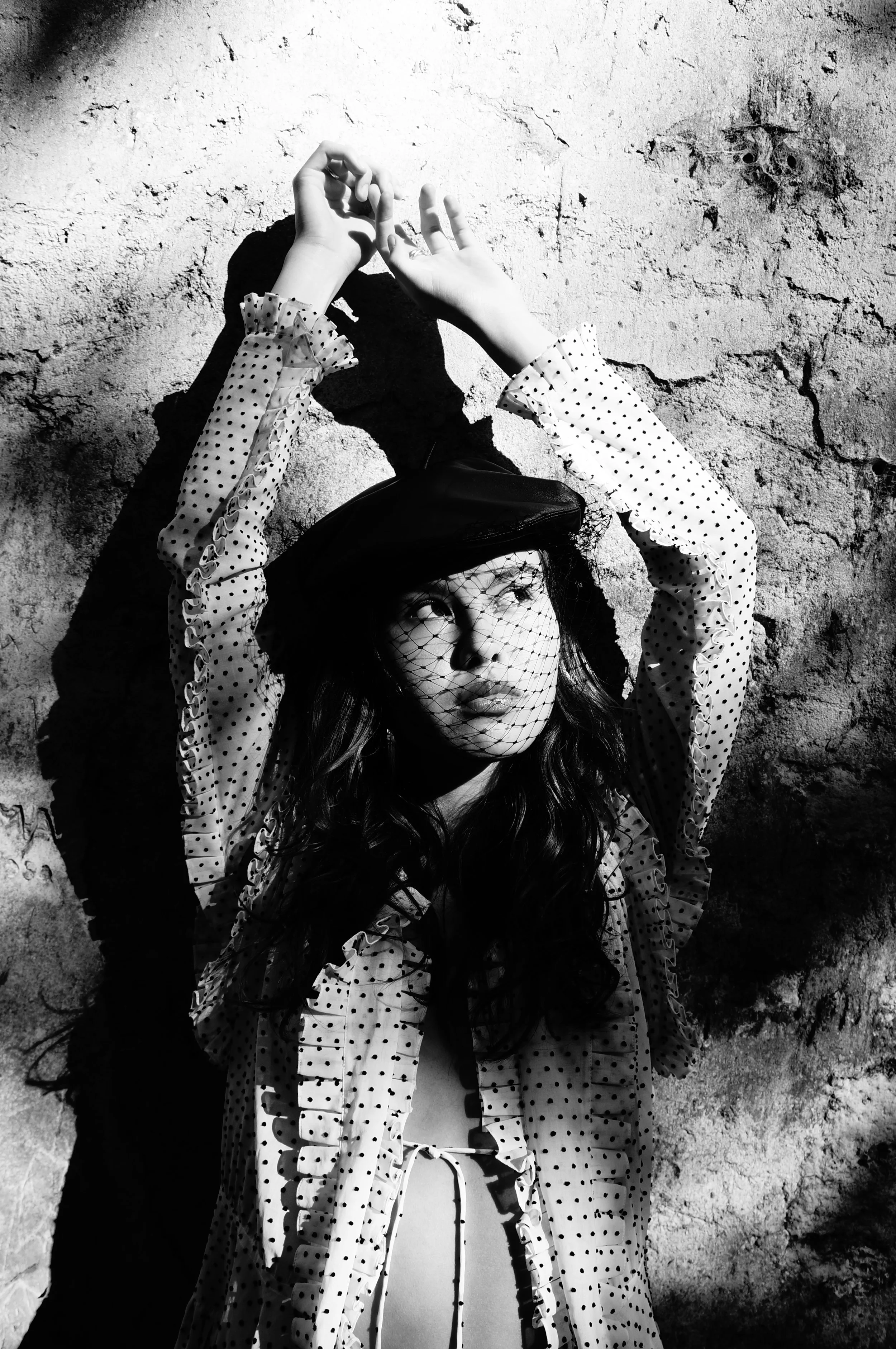 Black and white photo of a woman with wavy hair wearing a polka-dot blouse, a hat with a veil, and a tie-front top, standing against a textured wall with her hands raised above her head.