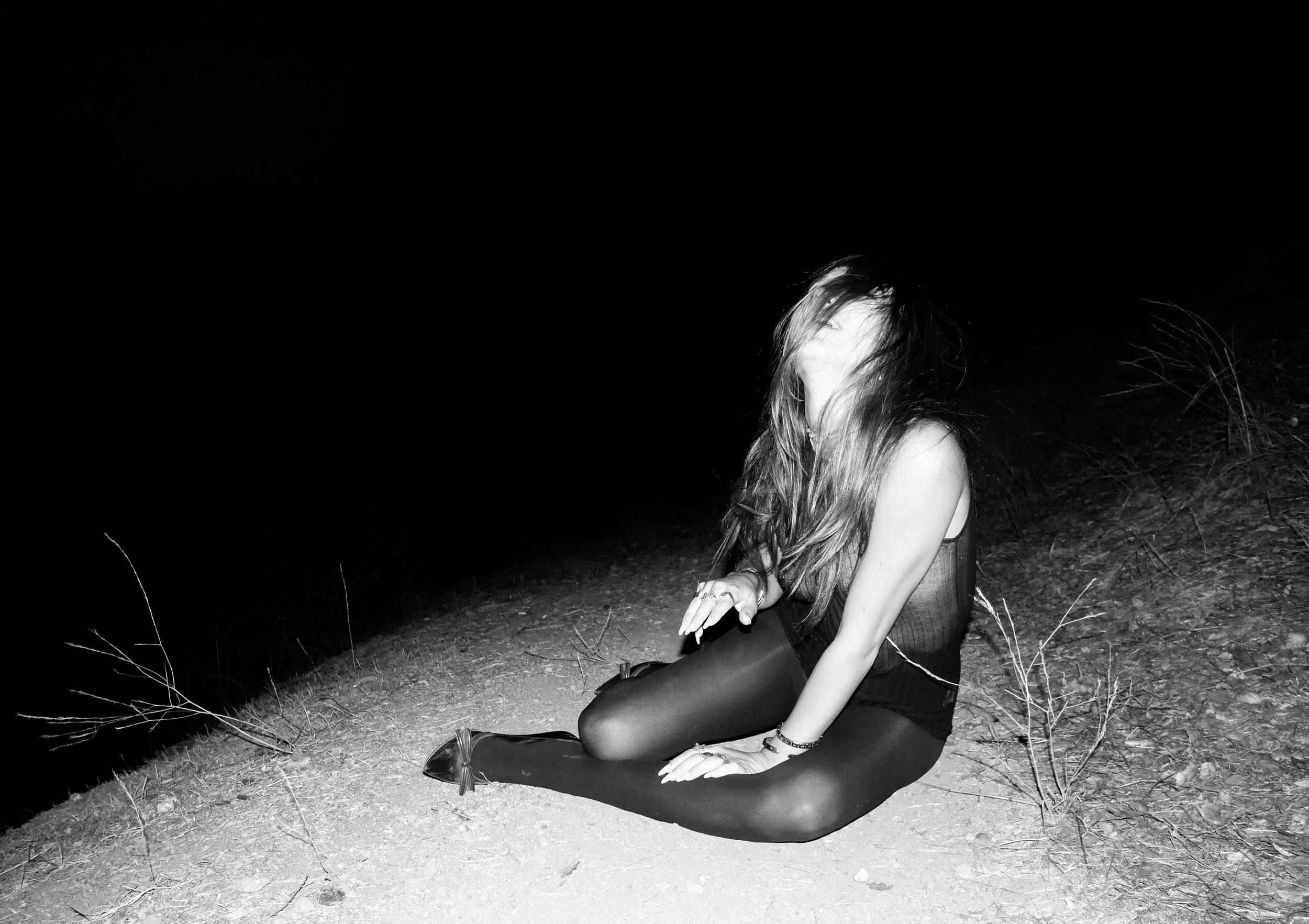 A woman sits on dirt with her head down and hair covering her face, at night, with dark background.