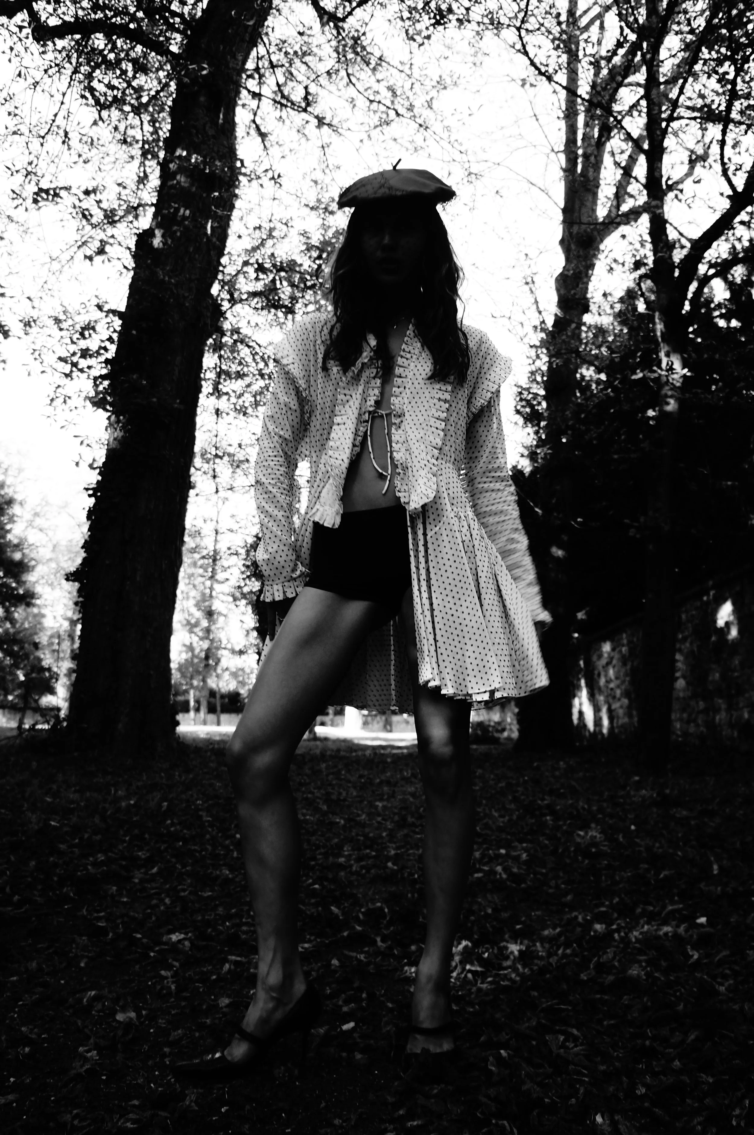 A woman standing outdoors near trees, dressed in a polka dot coat, shorts, high heels, and a hat, with a background of tall trees and overcast sky.