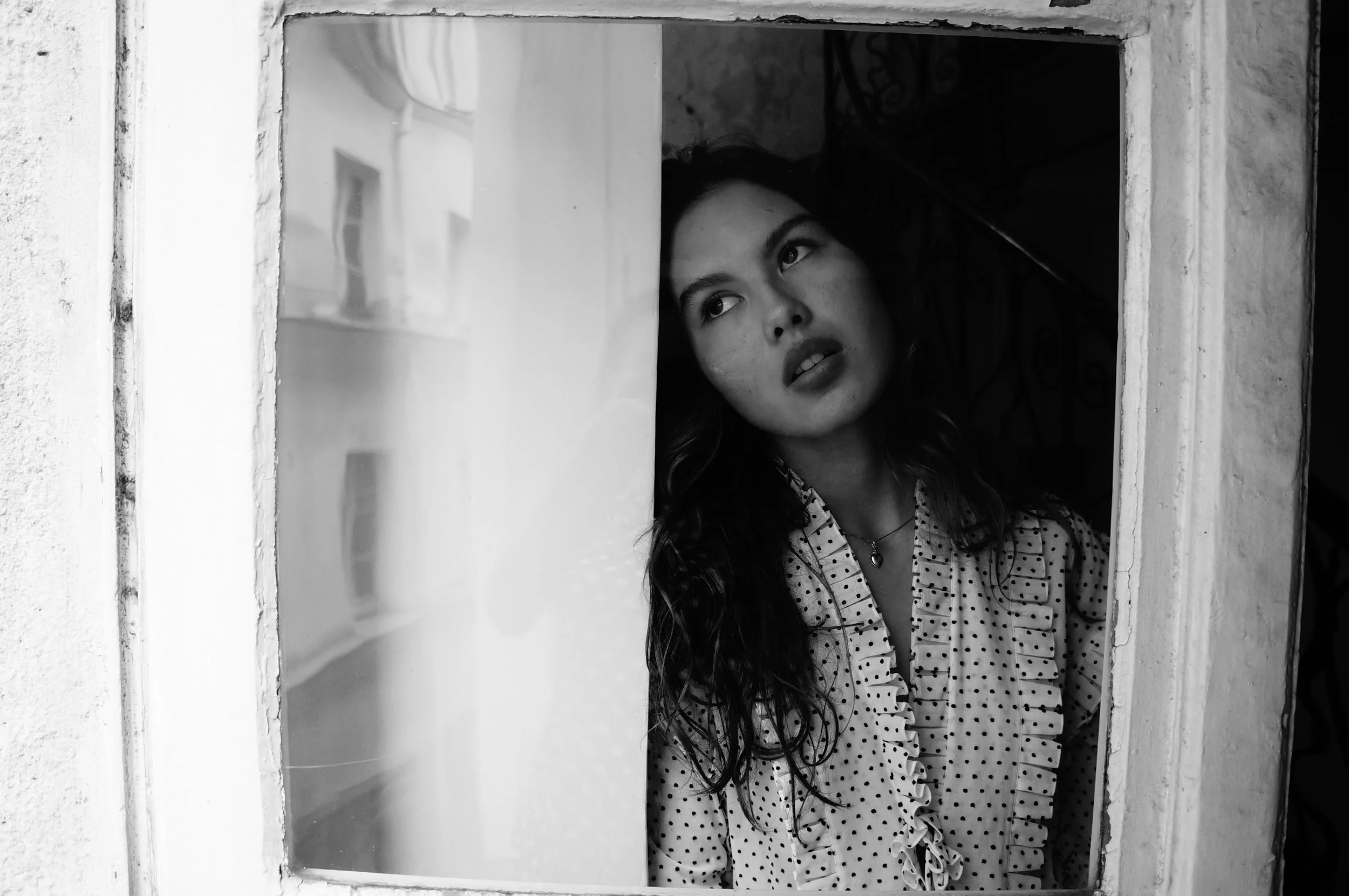 A woman with long hair and a polka dot blouse looking through a window in a black and white photo.