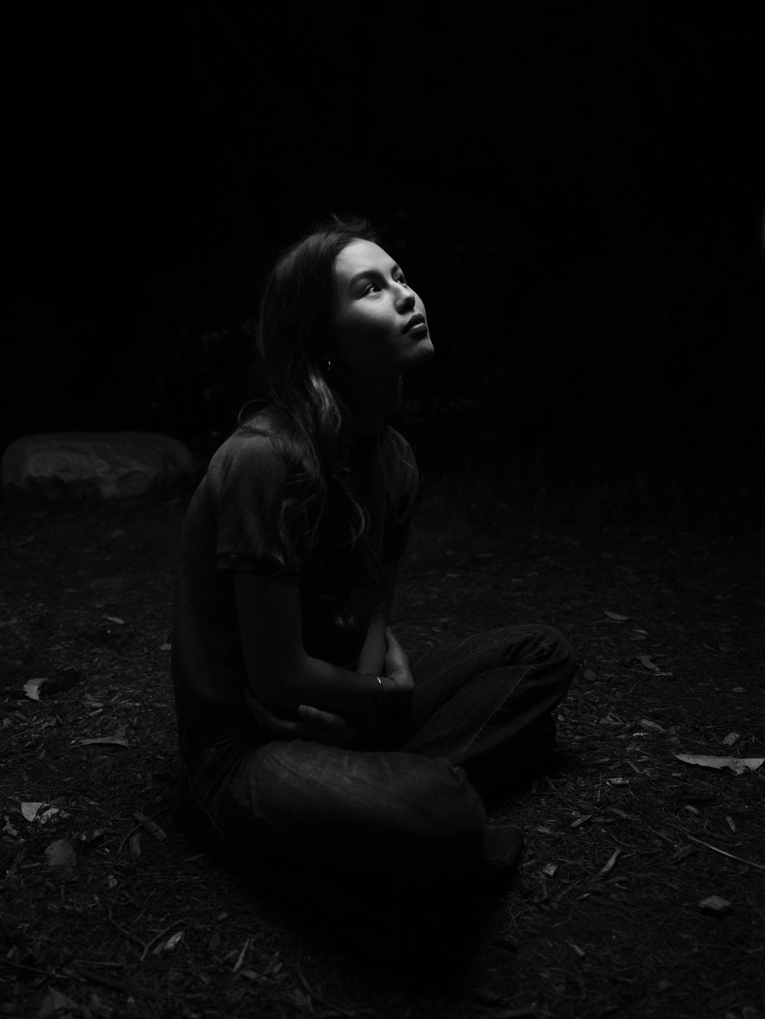 A woman sitting on the ground at night, looking upwards, illuminated by a soft light, with trees and rocks in the background.
