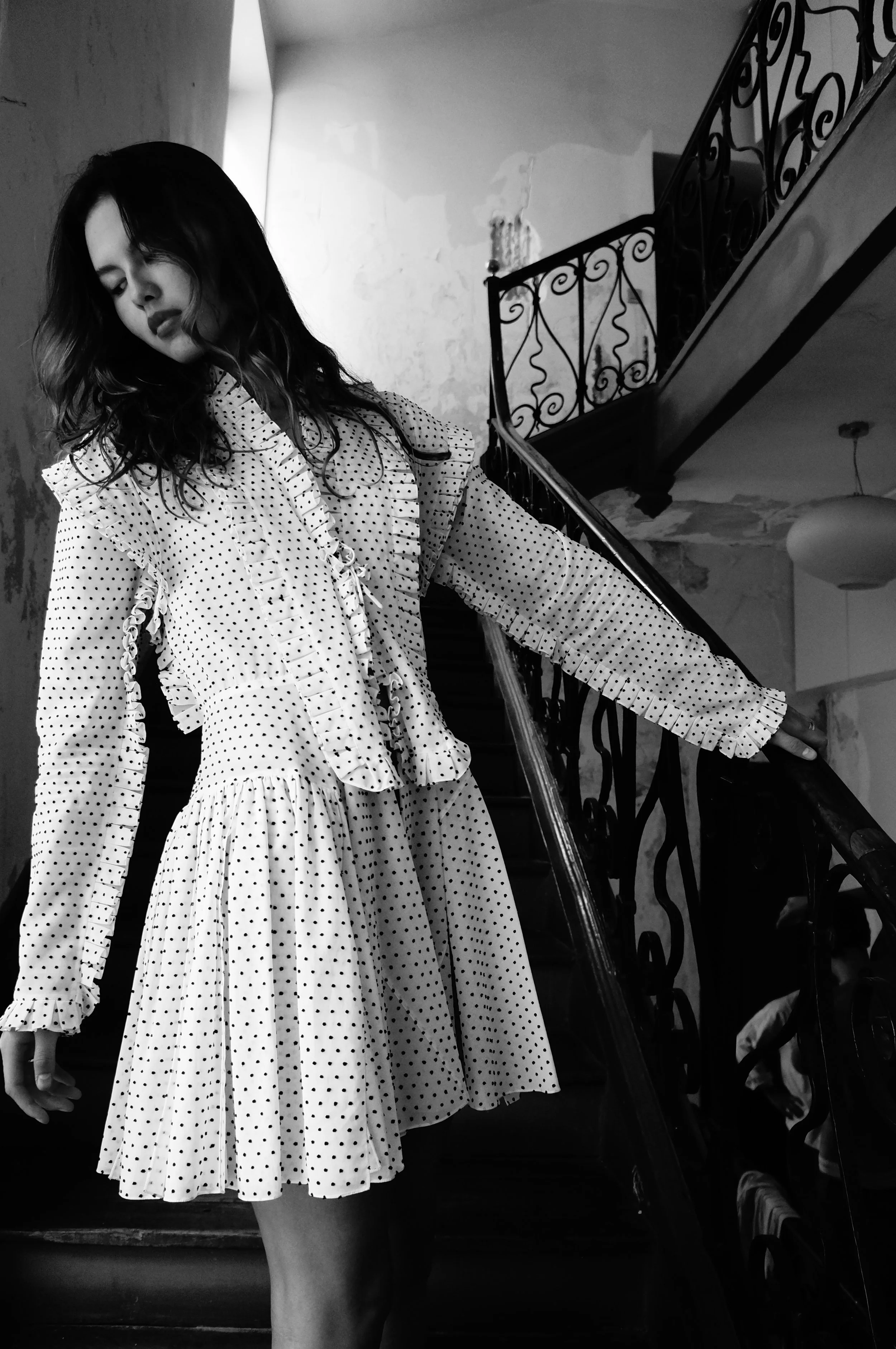 A woman with wavy hair wearing a polka dot dress standing on a staircase, with her eyes closed and hand resting on the railing.