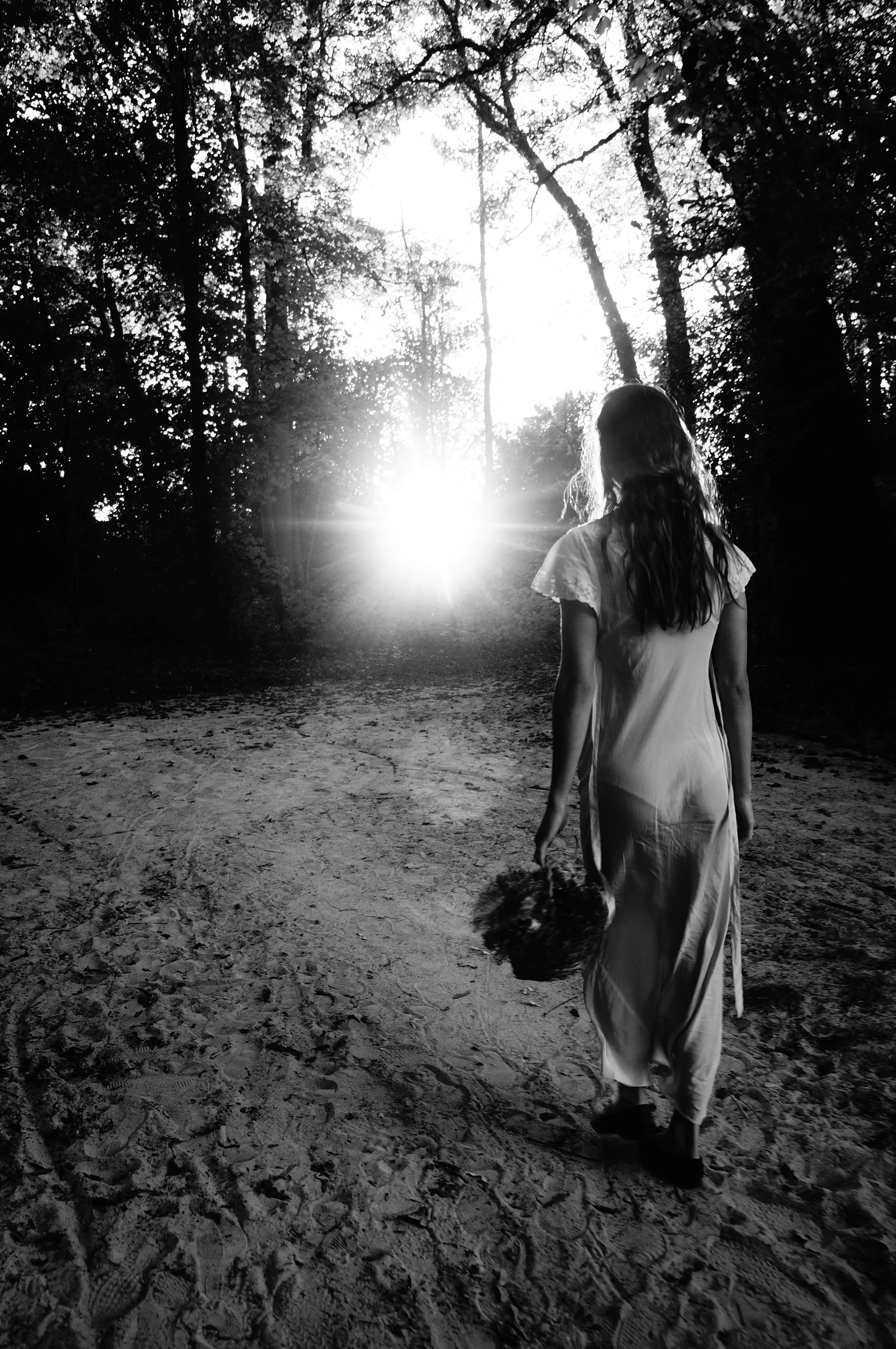 Black and white photo of a girl walking away on a wooded dirt path, holding a bouquet, with the sun setting or rising in the background.
