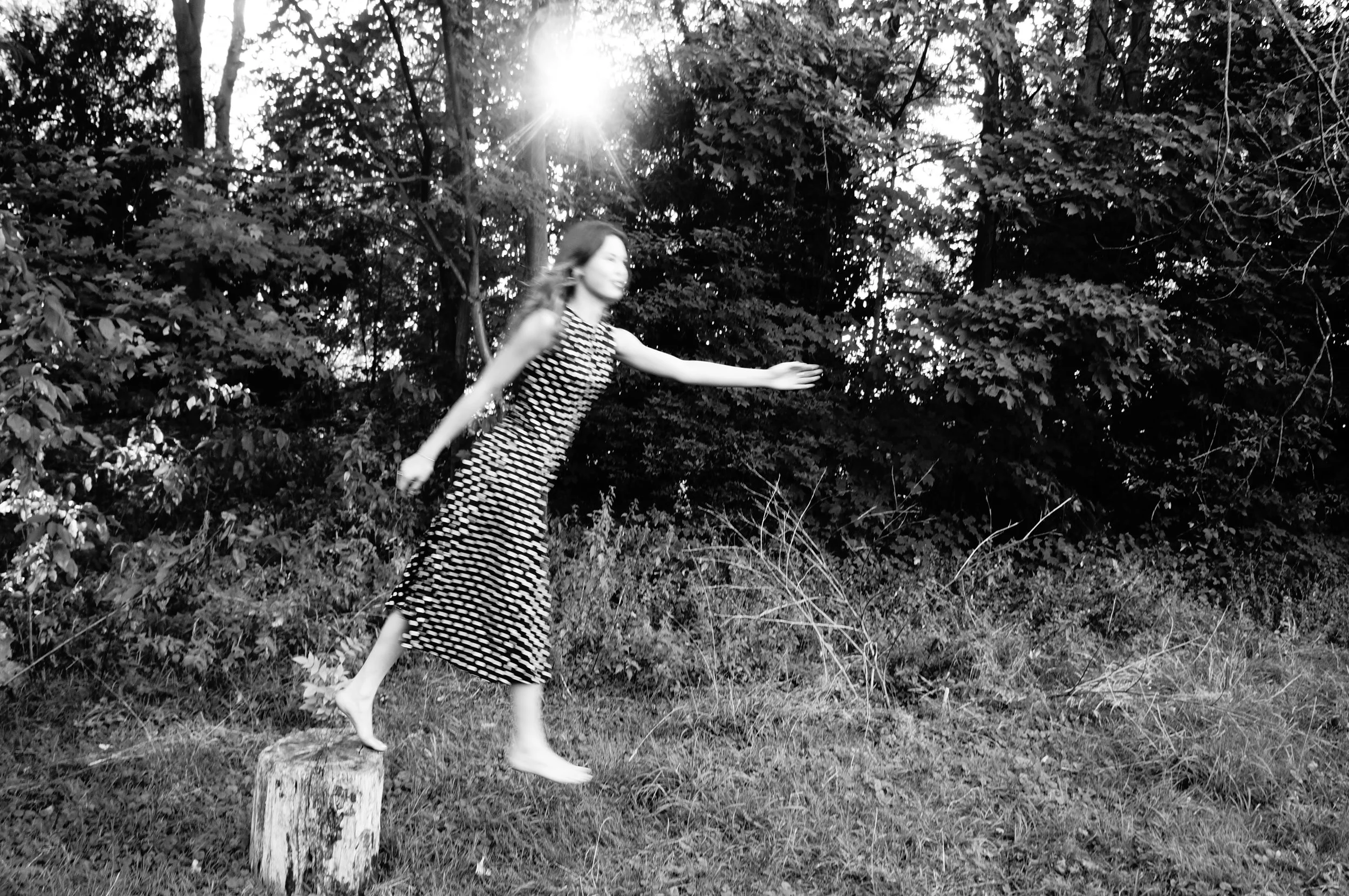 A woman in a patterned dress balancing on a tree stump in an outdoor wooded area at sunset, with sunlight filtering through the trees.