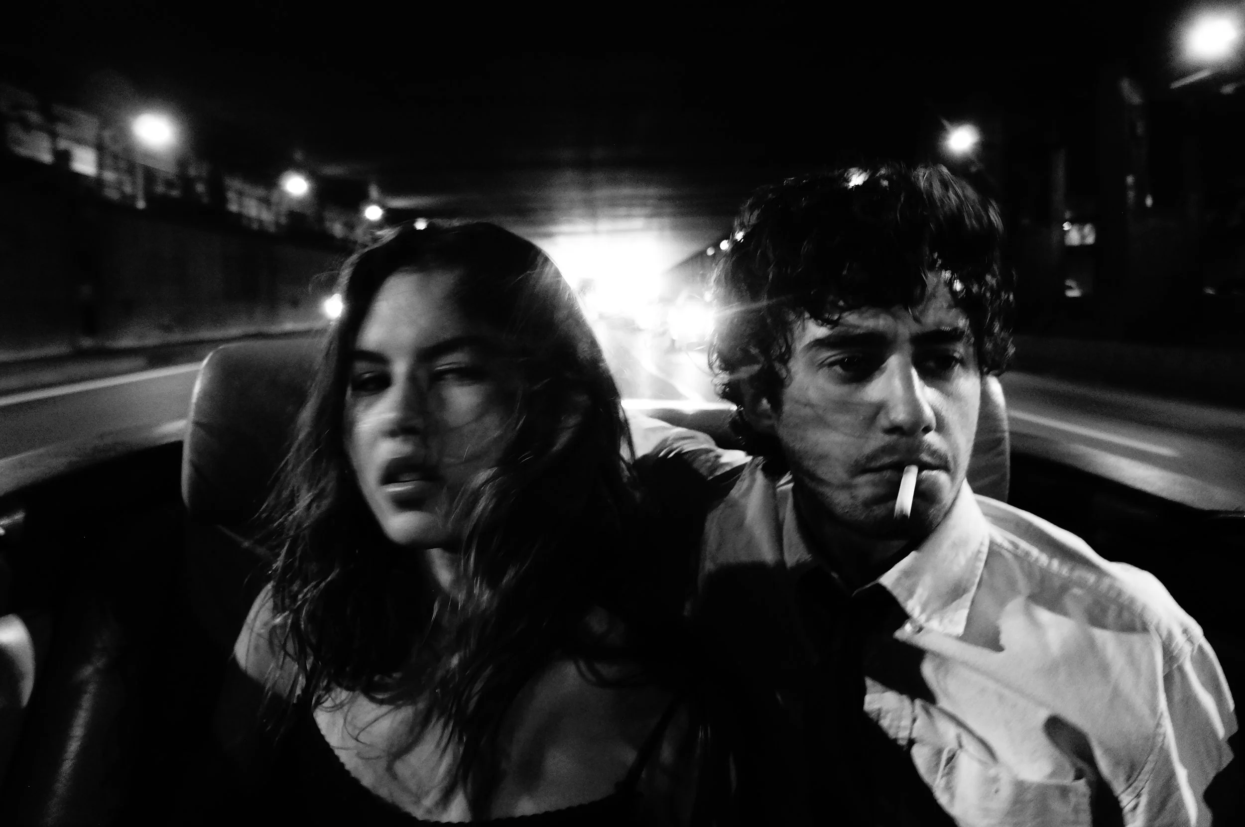 A black and white photo of a woman and a man sitting together in the back seat of a moving car at night, with city lights flashing in the background. The woman has long hair and is winking, while the man has curly hair, a cigarette in his mouth, and 
