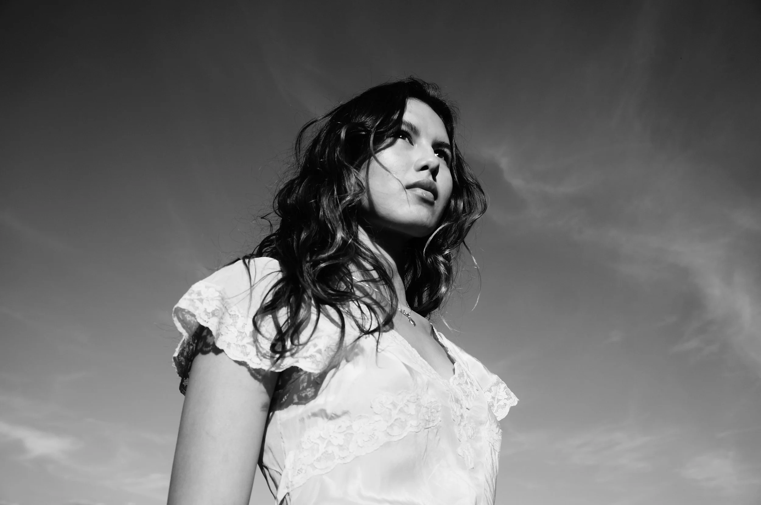 Black and white photo of a woman with wavy hair looking upwards, wearing a white lace top, against a partly cloudy sky.