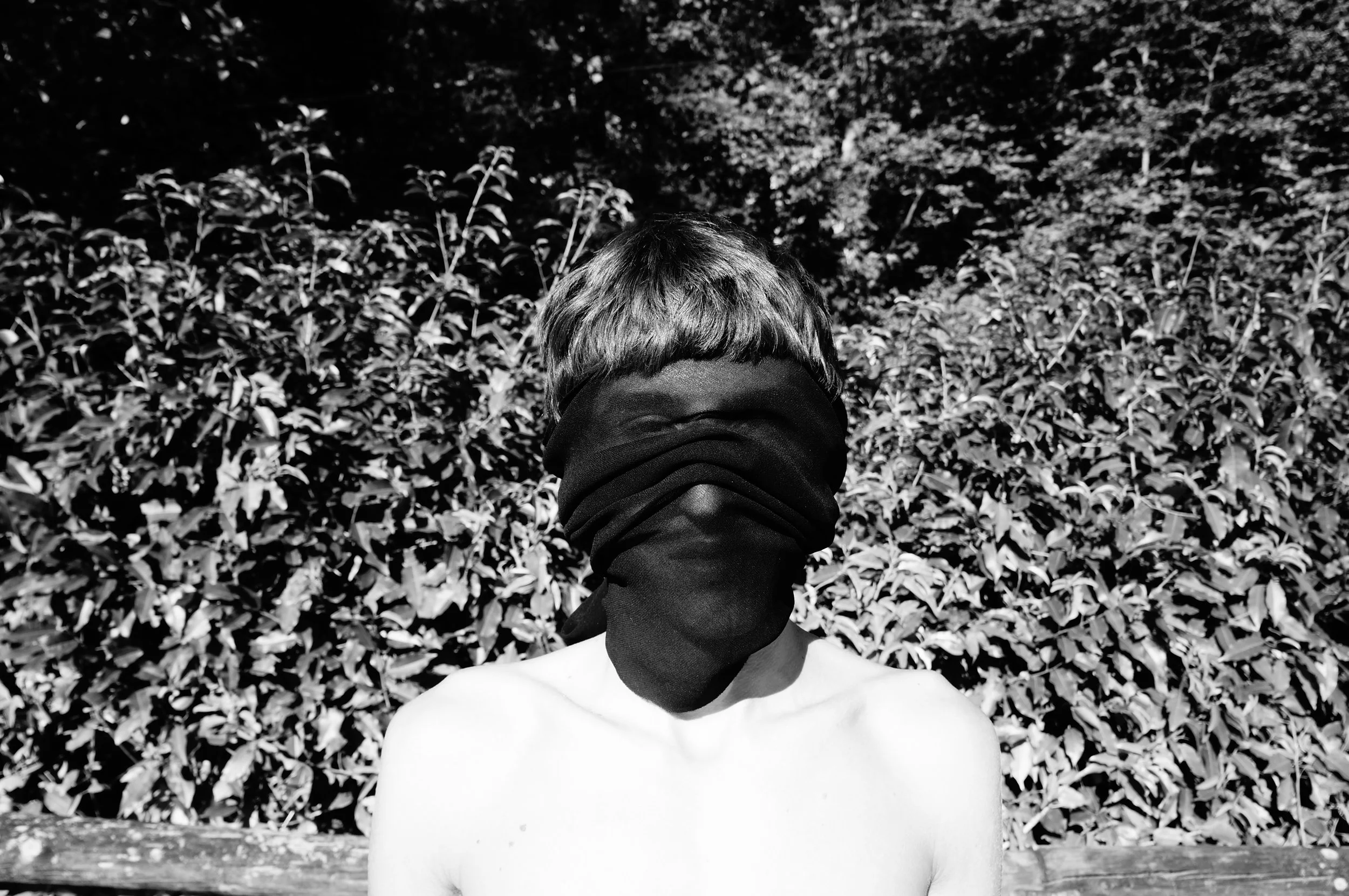 A shirtless person with a short haircut has their face covered by a black cloth, standing outdoors in front of dense foliage in black and white.