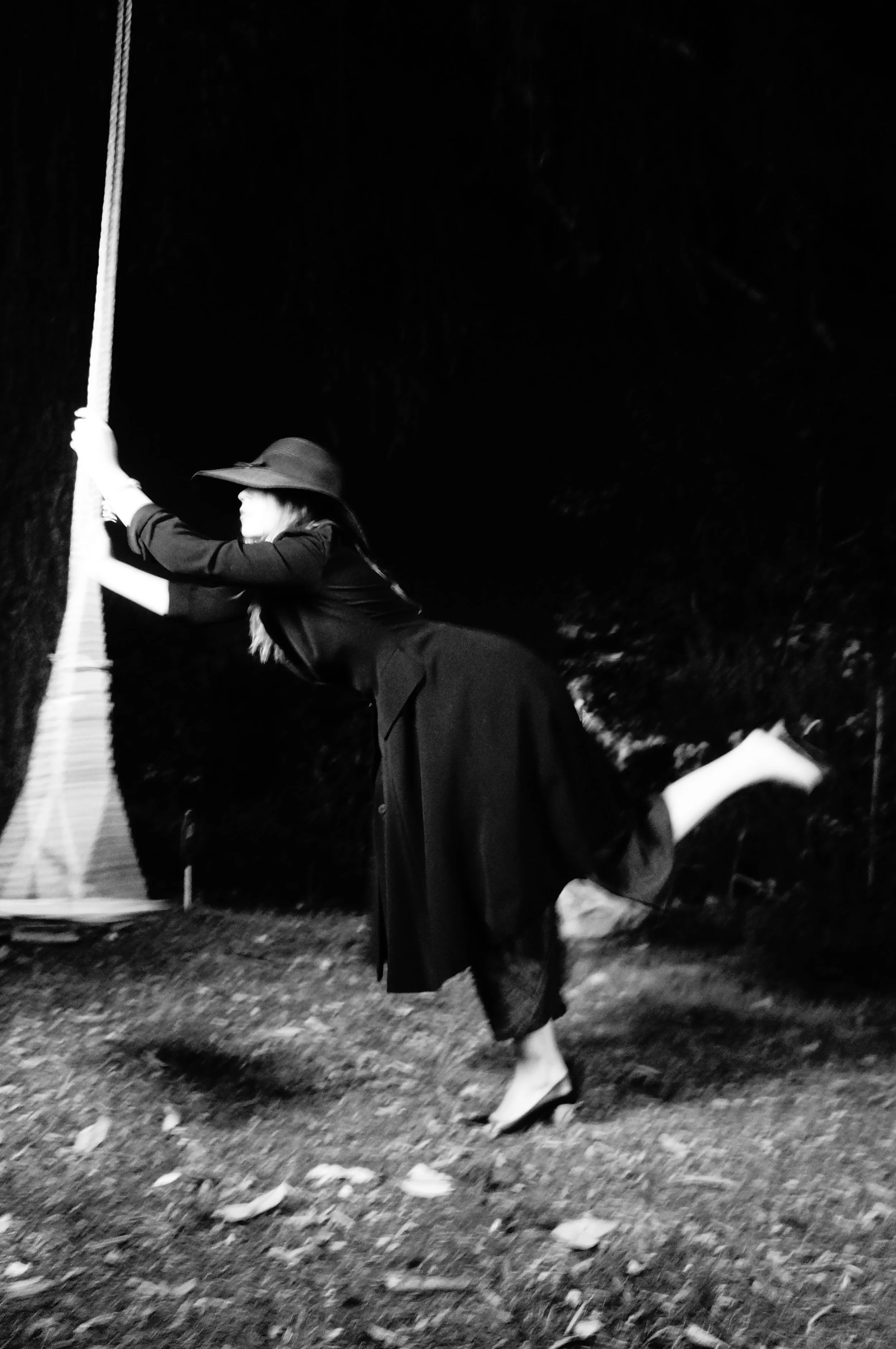 A woman in a black dress and a wide-brimmed hat is playfully pulling a rope at night, standing barefoot on a grassy and leaf-strewn ground.