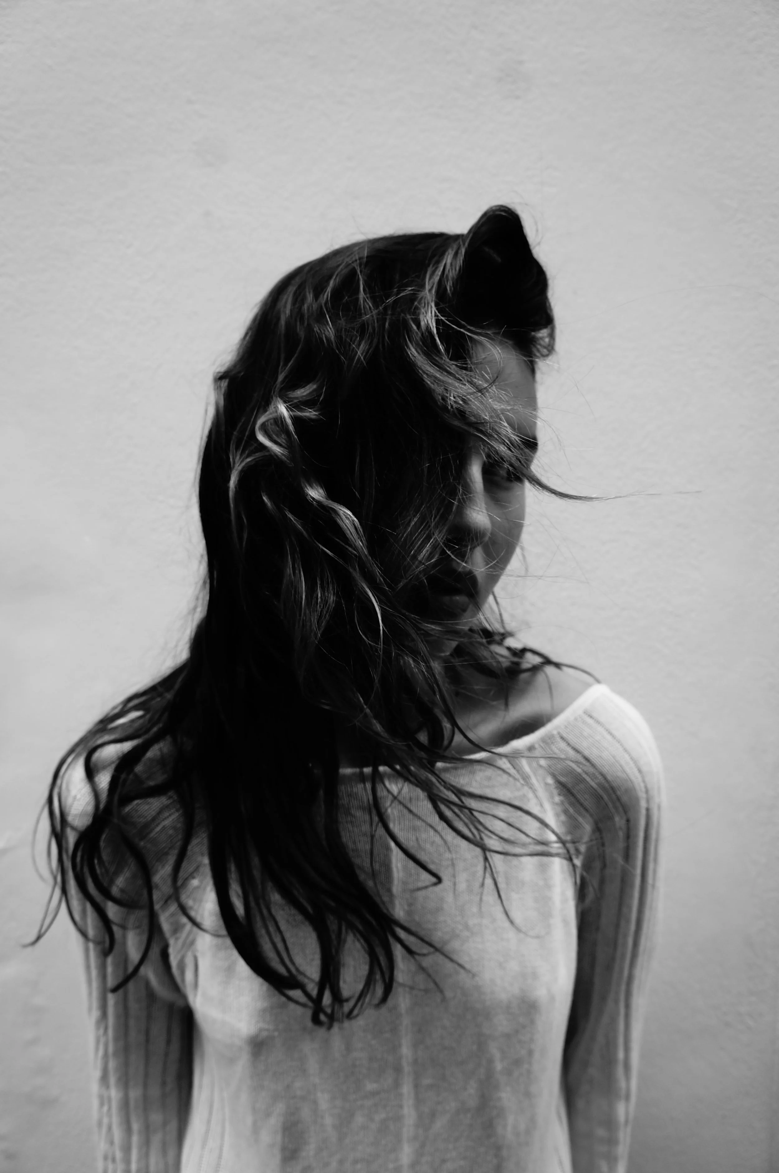 A black-and-white photo of a woman with wet, wavy hair partially covering her face, looking down slightly, wearing a light-colored, long-sleeve shirt, standing against a plain wall.