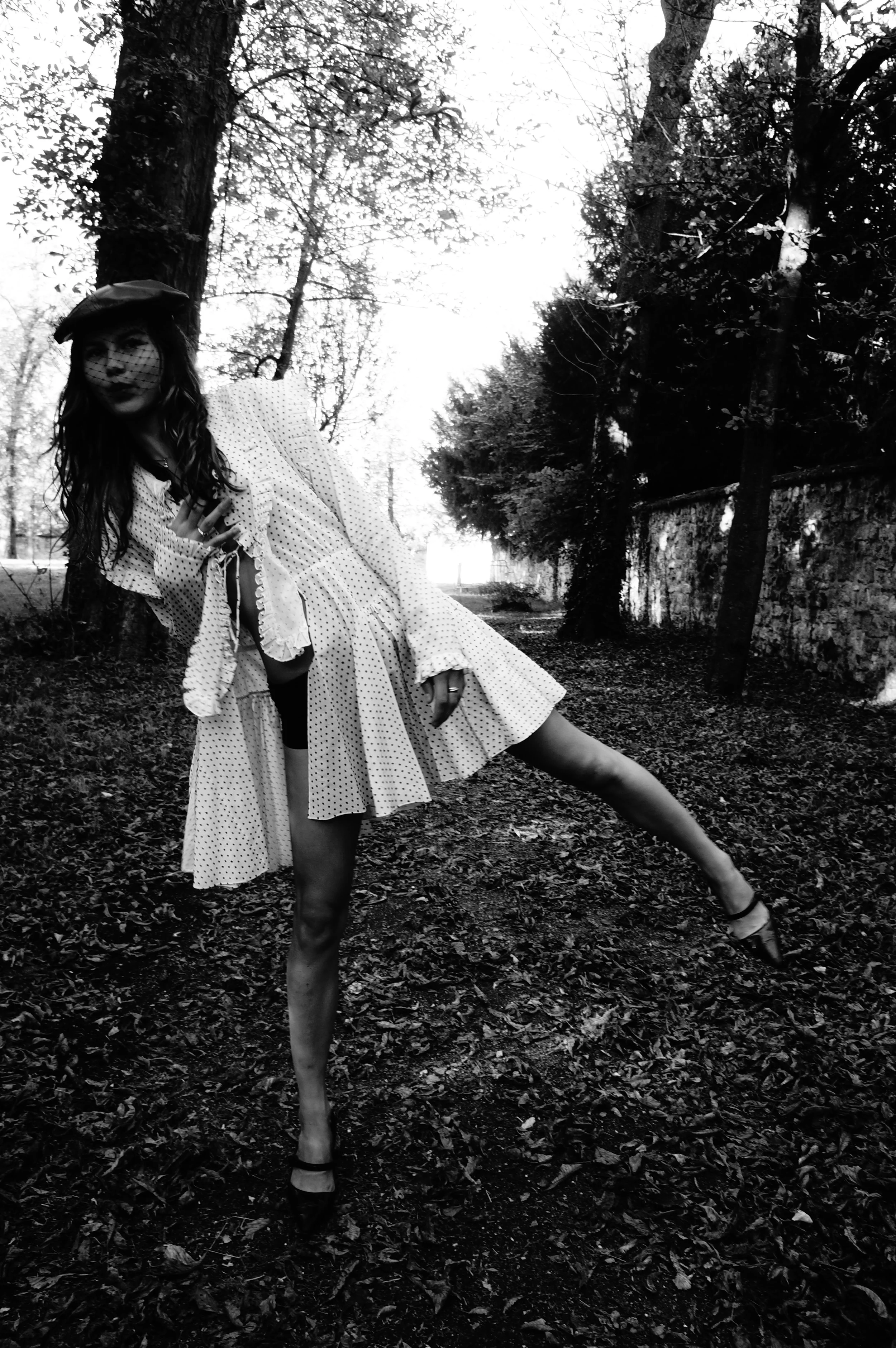 A woman wearing a polka dot dress, hat, and veil posing outdoors among trees and fallen leaves on the ground.