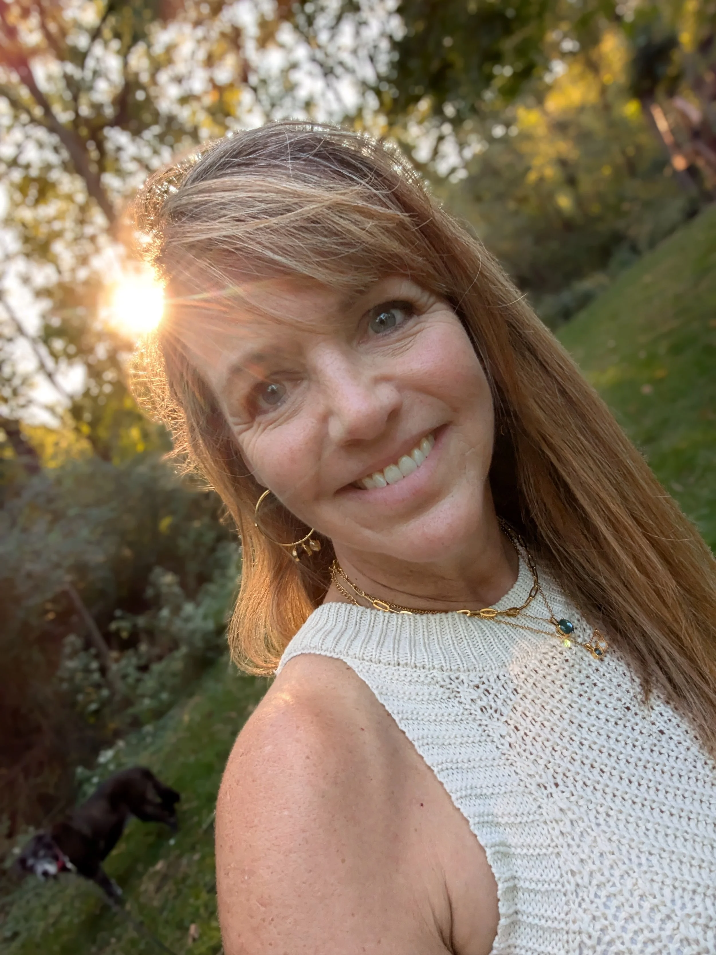 A smiling woman with long reddish-brown hair takes a selfie outdoors during sunset, with a dog in the background and trees with yellow leaves.