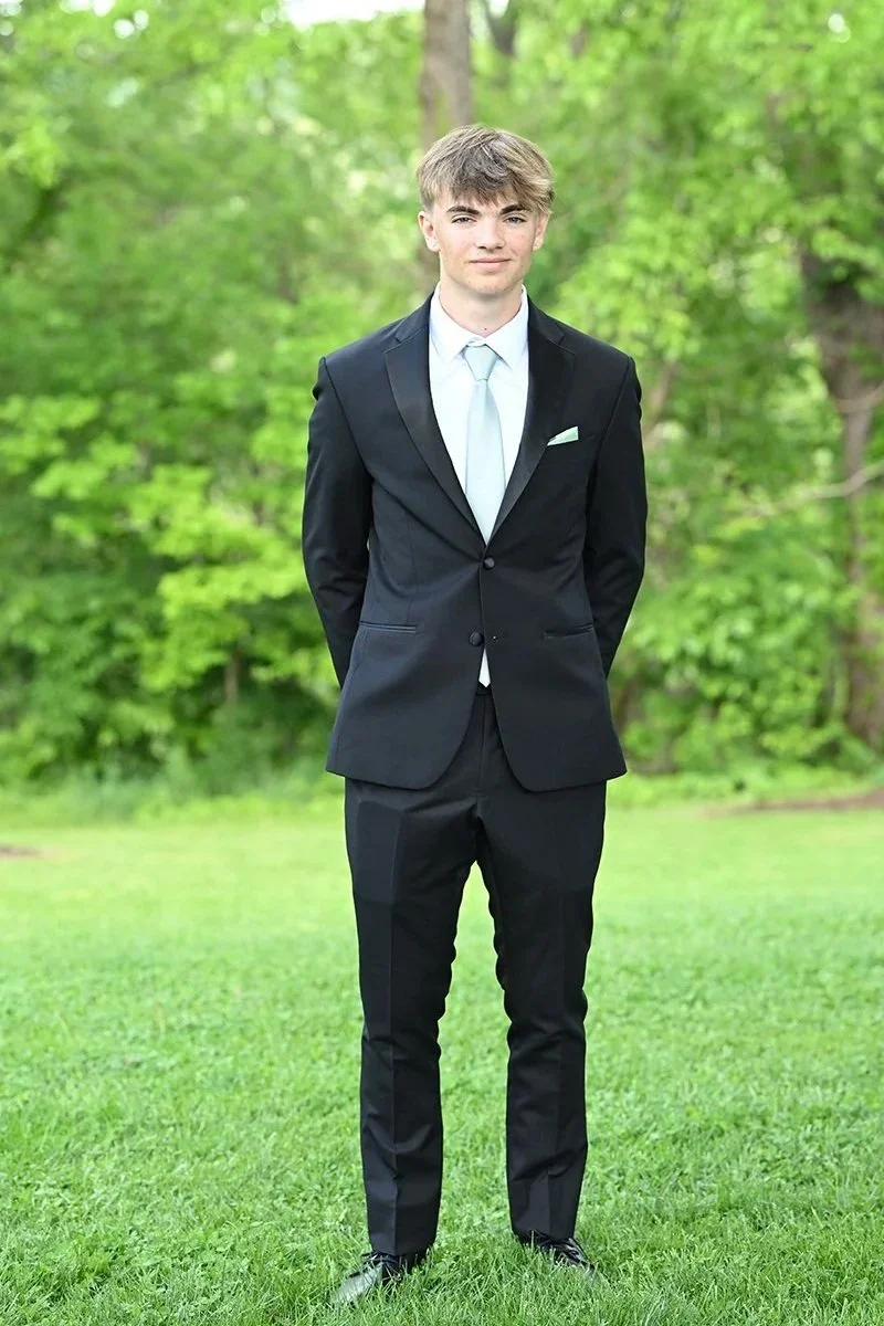 Young man in a black suit with a light blue dress shirt and tie standing outdoors on grass with green trees in the background.