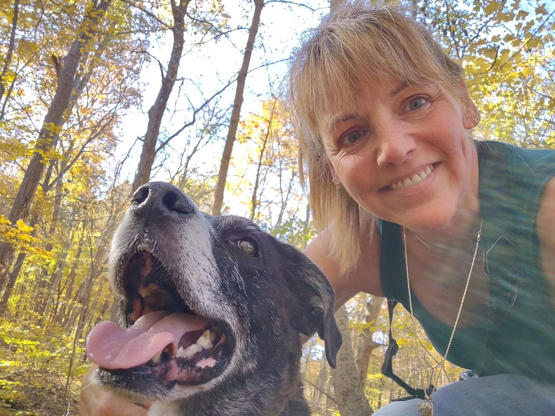 A woman smiling with a black and white dog in a forest during fall.