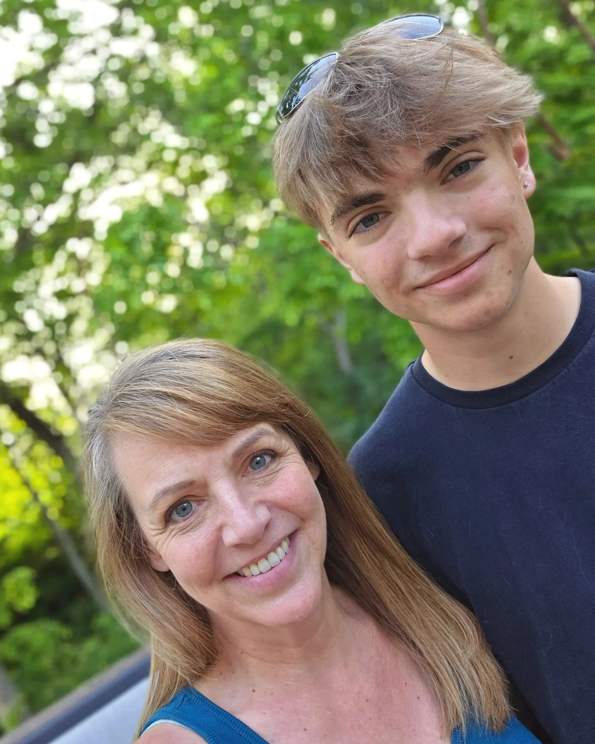 Close-up of a woman with long red hair and blue eyes smiling outdoors with green foliage in the background, and a young man with light brown hair and sunglasses on his head smiling next to her.