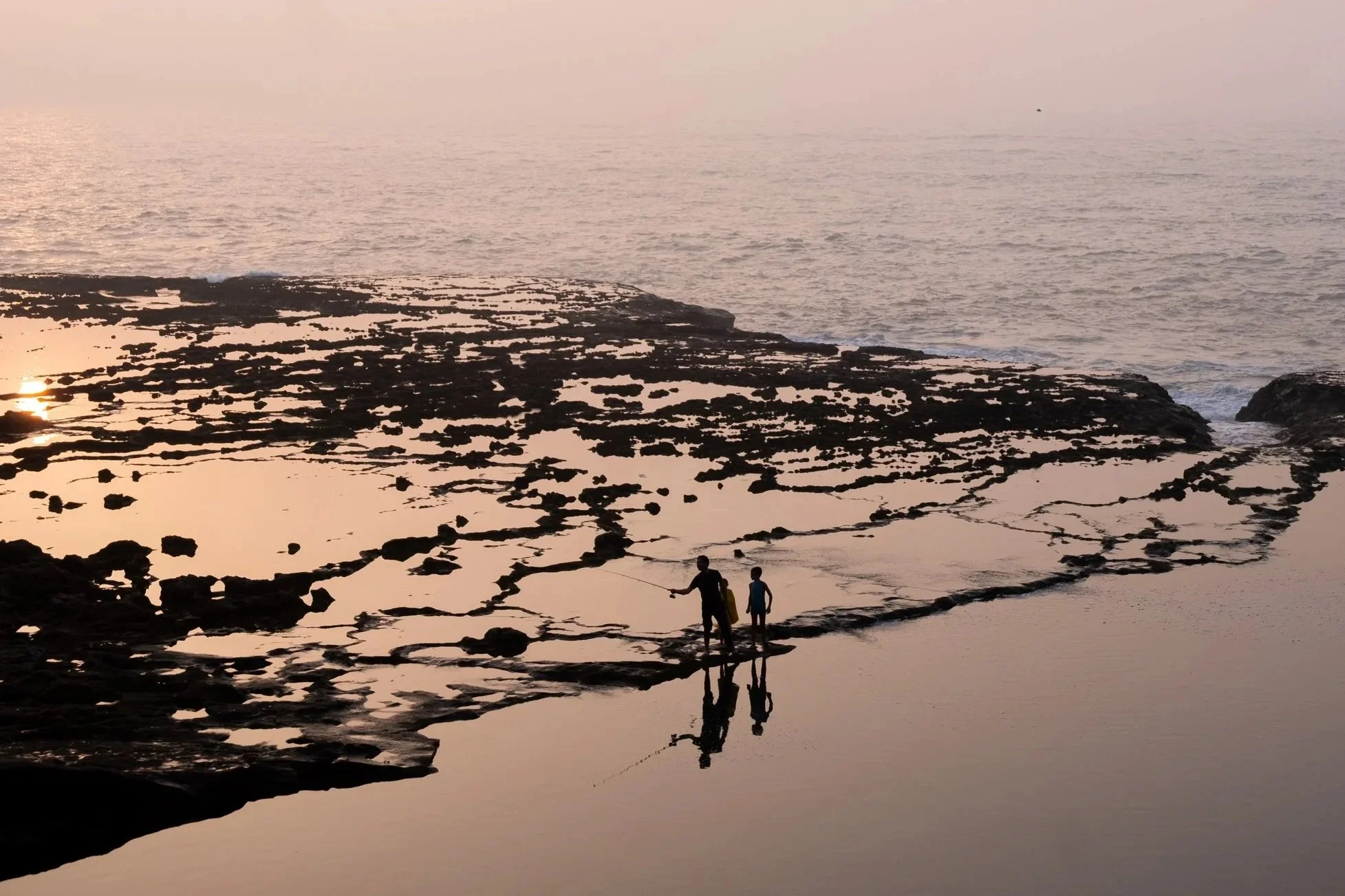 Family silhouette along the rocky Atlantic shoreline and tide pools at sunset in Essaouira, Morocco.
