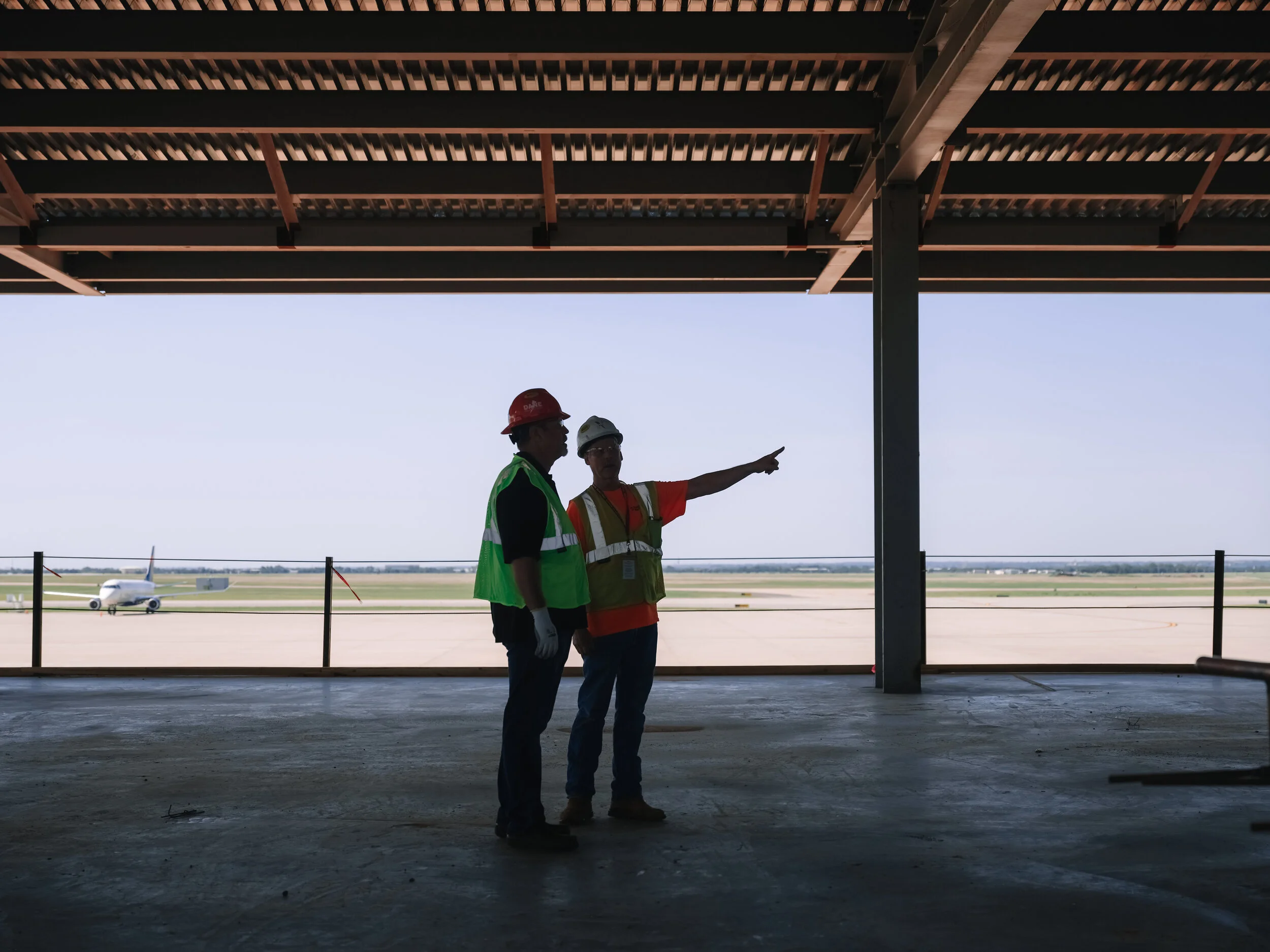 Two construction workers wearing safety vests and helmets standing inside an airport terminal, looking out at an airplane on the runway, with one worker pointing towards the plane.