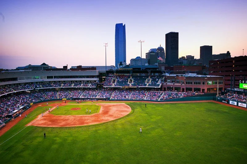 A baseball stadium filled with spectators, set against a city skyline at sunset with tall skyscrapers.
