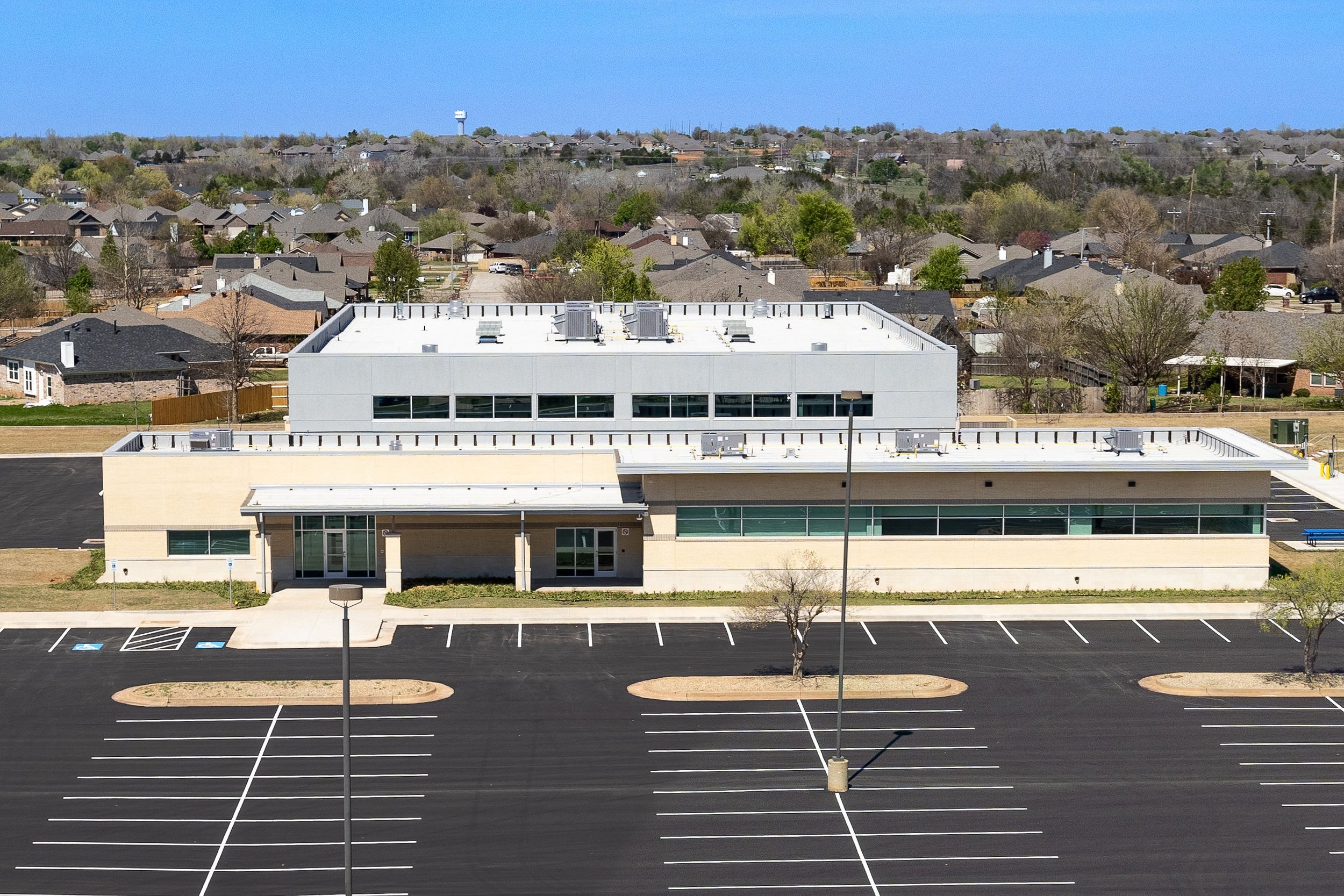 An empty parking lot in front of a modern, single-story building with glass windows and a light-colored exterior, with a second-story structure on top and a background of residential houses and trees.