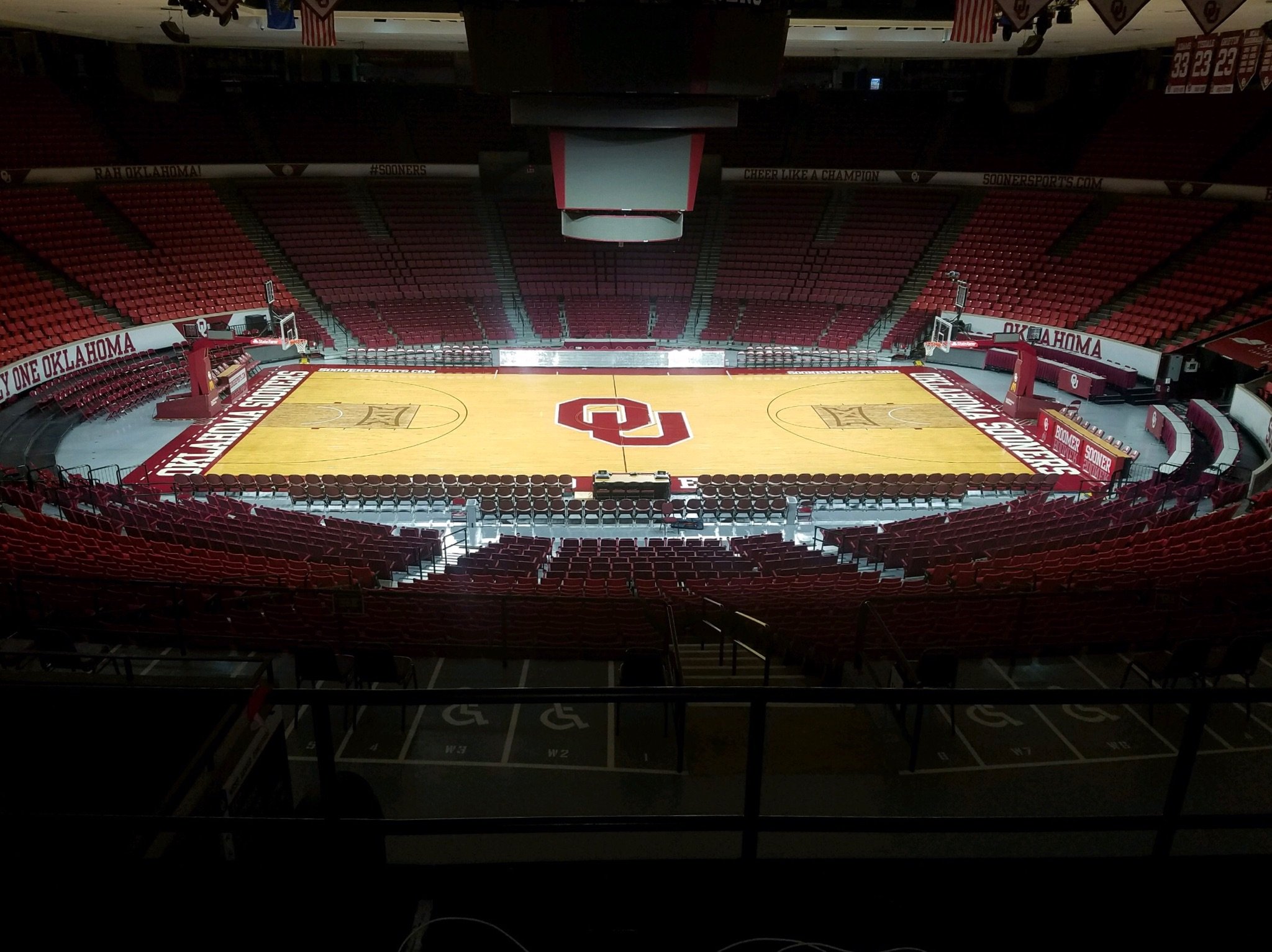 Interior view of a basketball arena with red seating, hardwood court, and the Oklahoma Sooners logo at the center. Scoreboards and banners are visible, with empty seats surrounding the court.