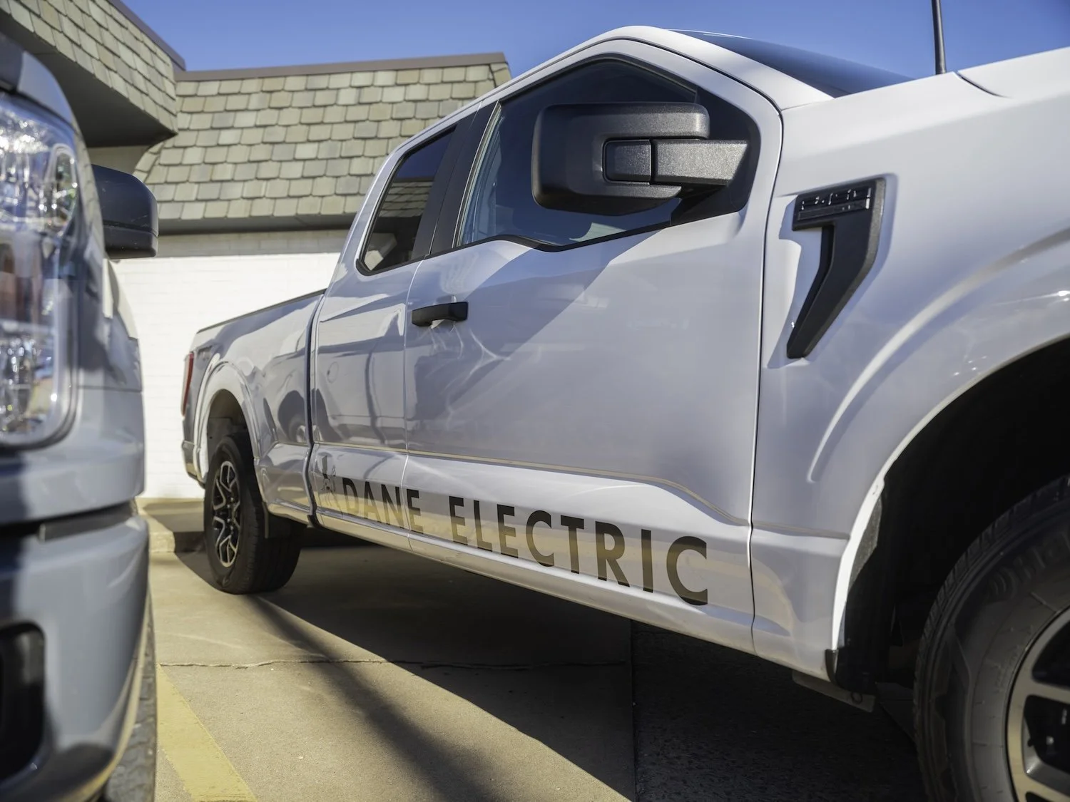 White electric pickup truck with 'DANE ELECTRIC' label parked in a parking lot, with a house and blue sky in background.